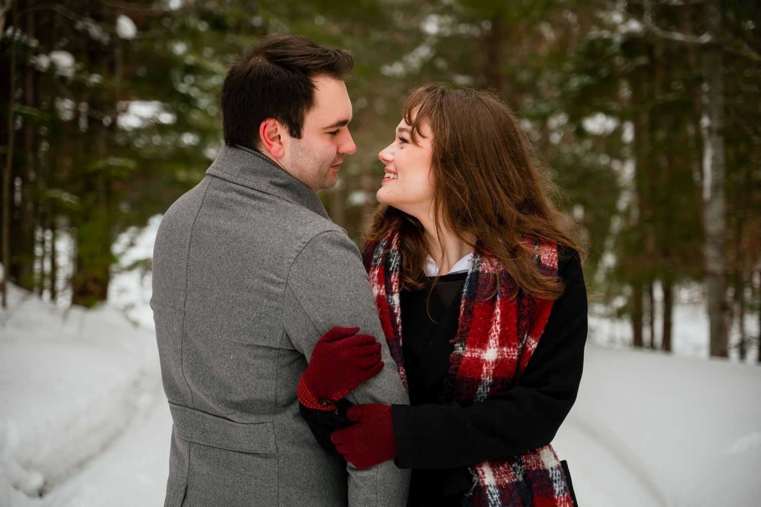 A couple in winter attire embraces romantically in a snowy forest setting with evergreen trees in the background.