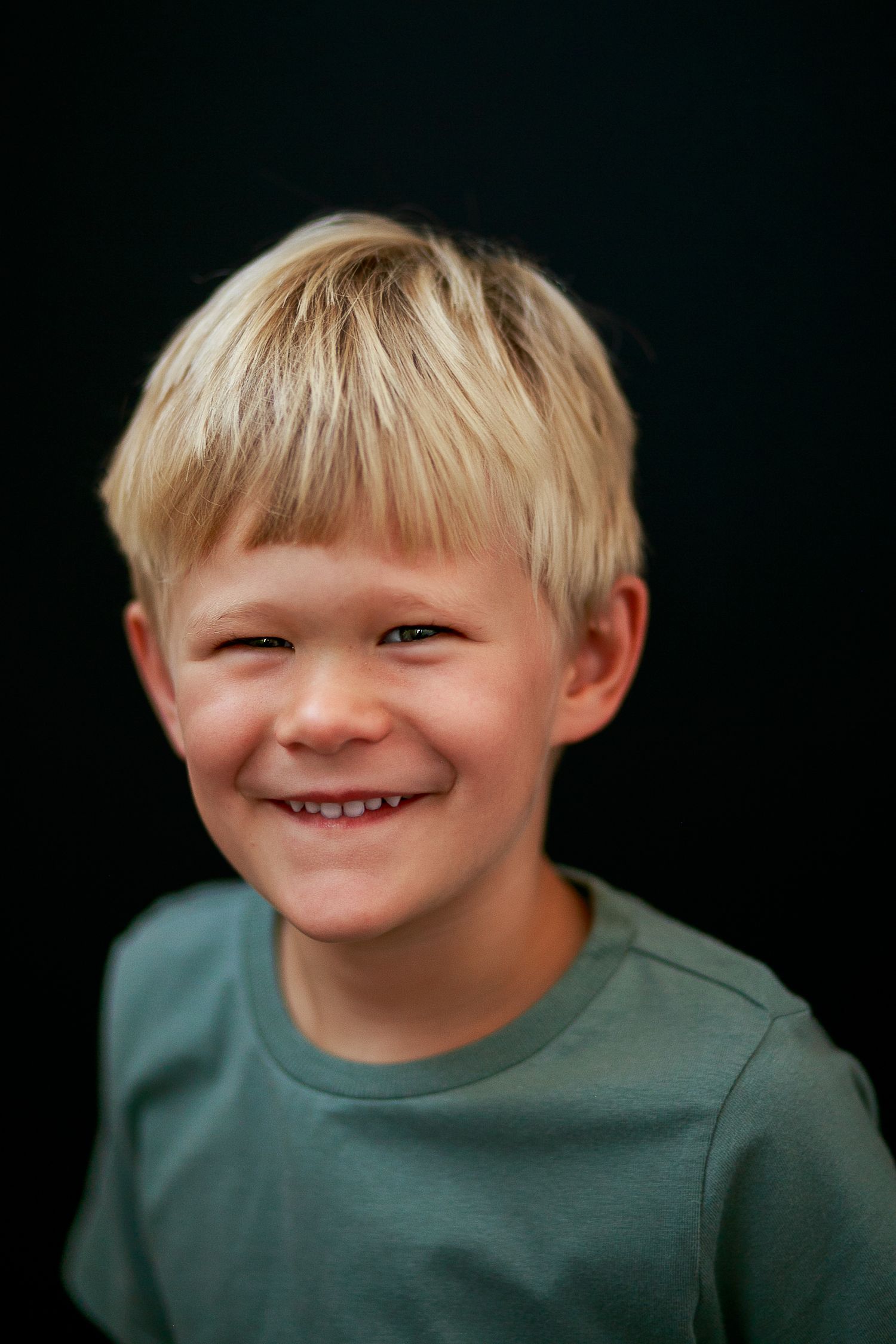 a child with blonde hair and a green shirt smiling against a dark background.