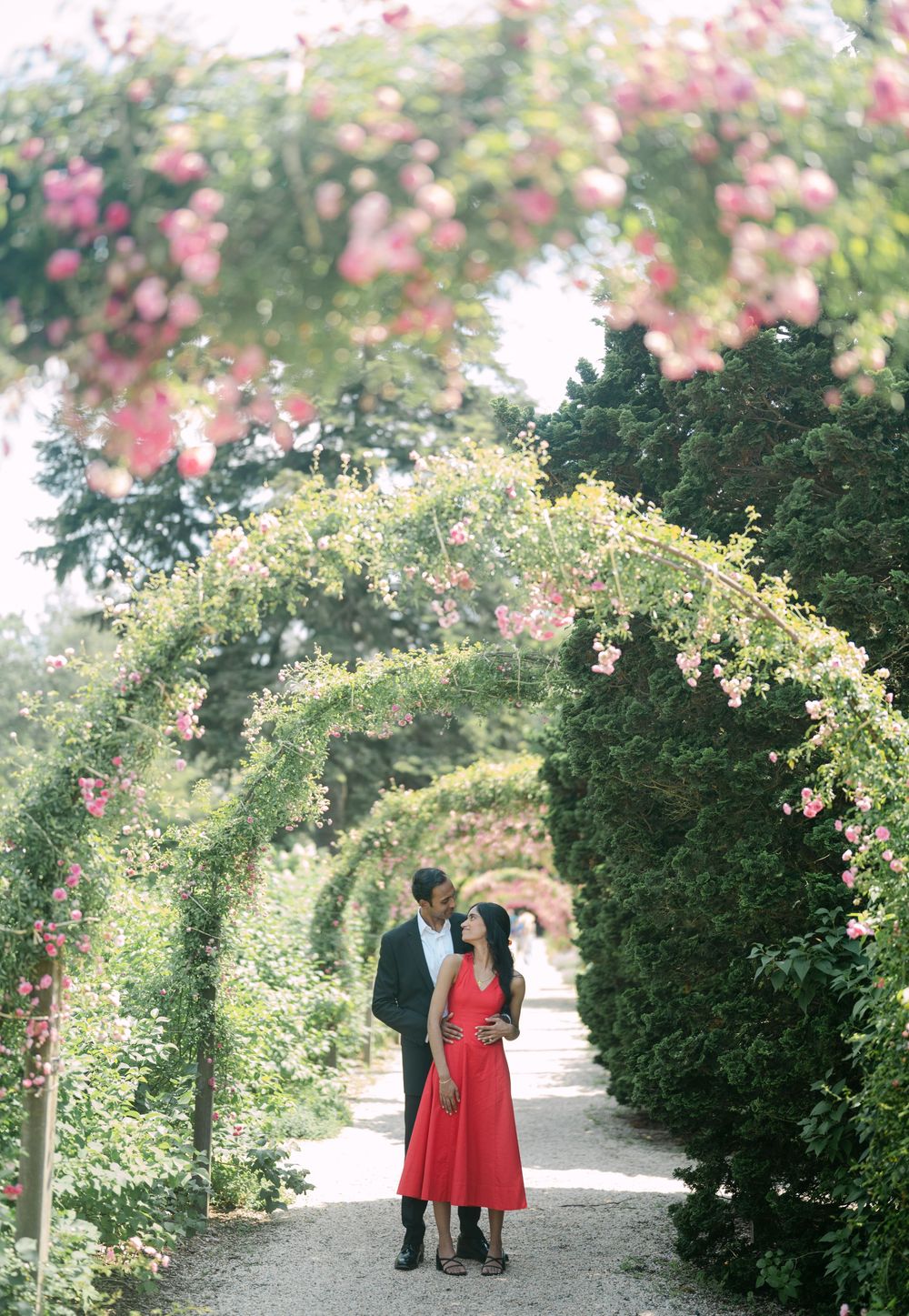 A couple stands beneath a romantic floral archway in a garden path surrounded by pink roses and greenery.