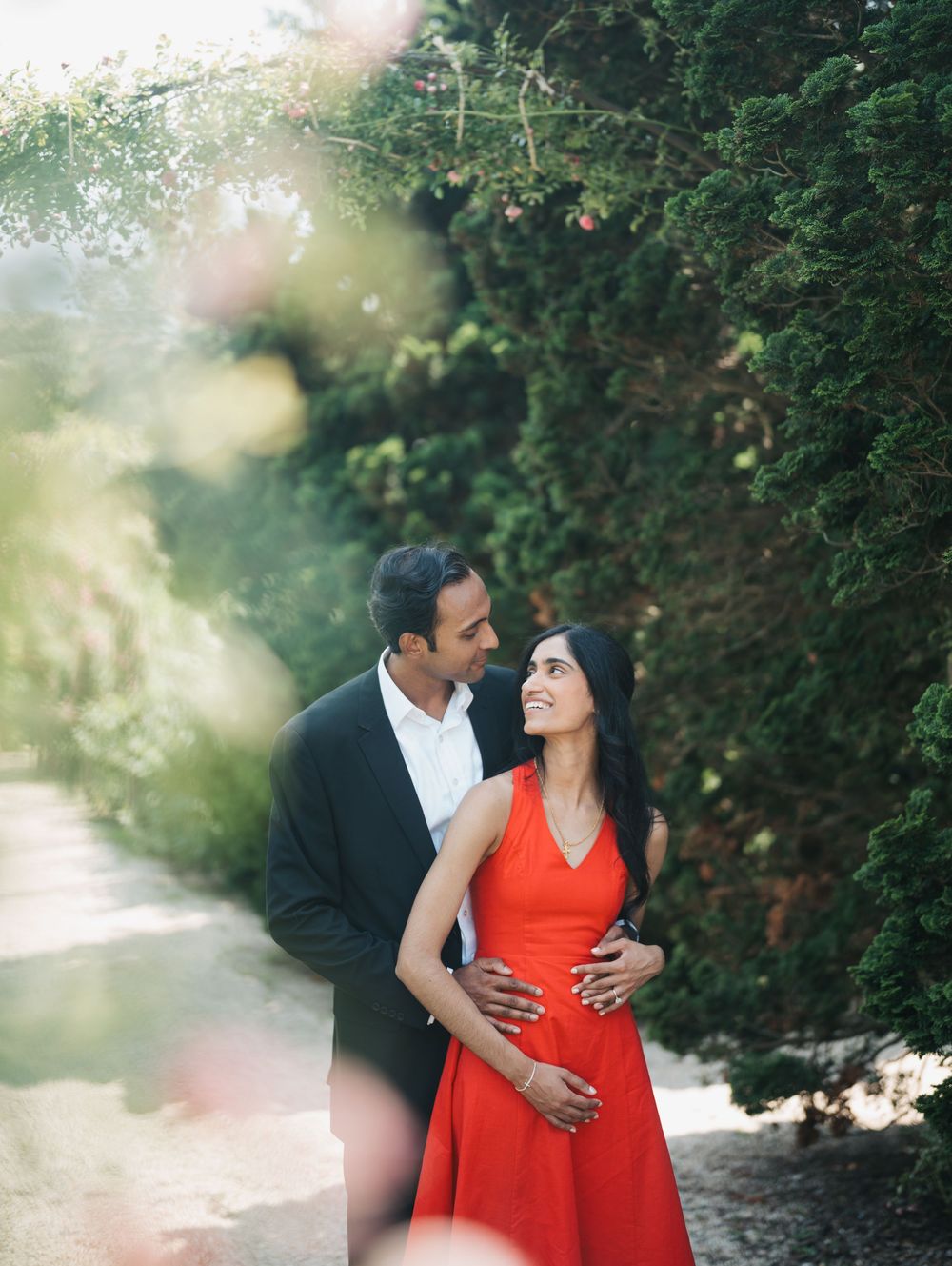 A couple shares a romantic moment in a lush garden setting with sunlight filtering through trees, the woman wearing a red dress.