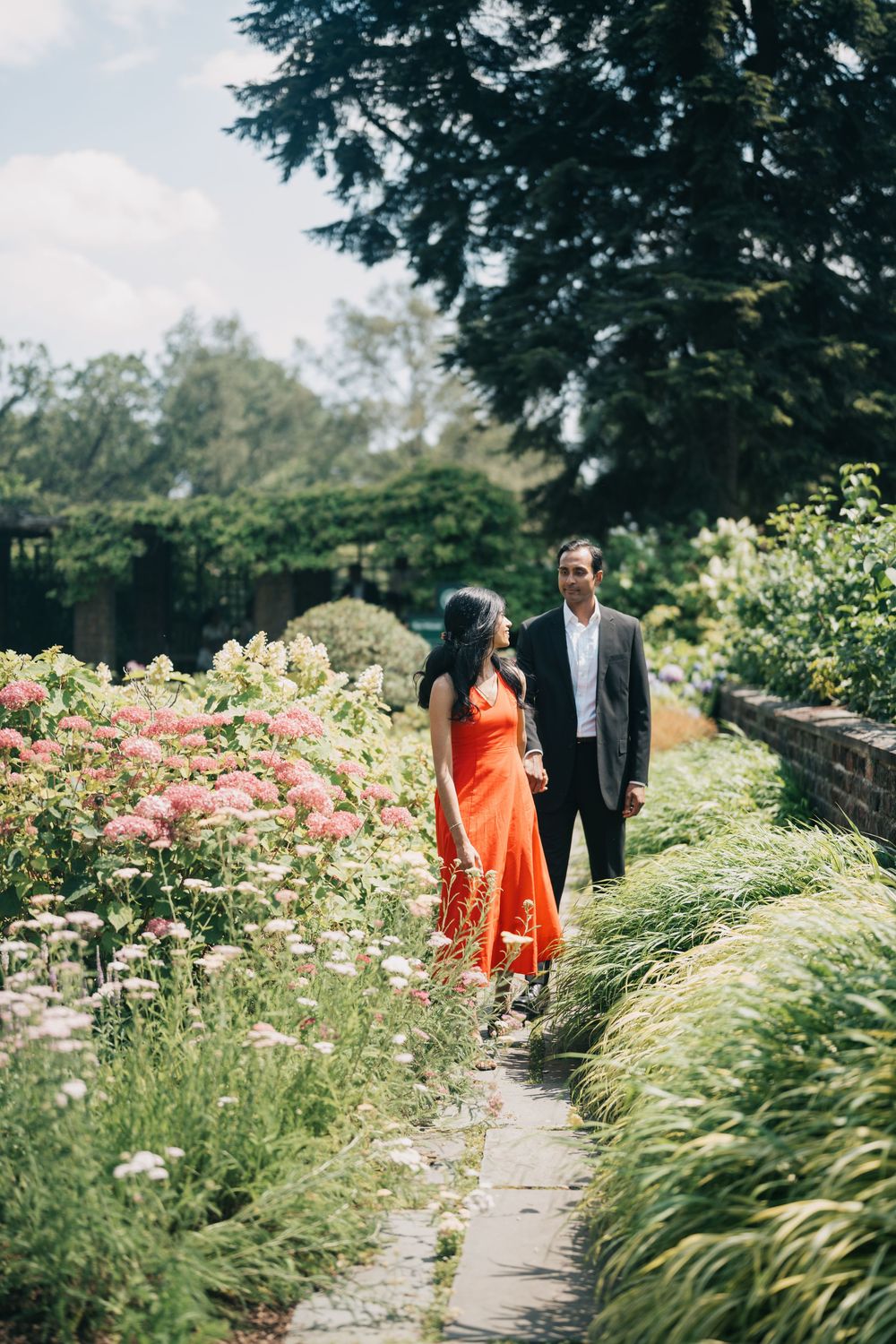 A couple walks along a garden path surrounded by blooming roses and ornamental grasses on a sunny summer day.