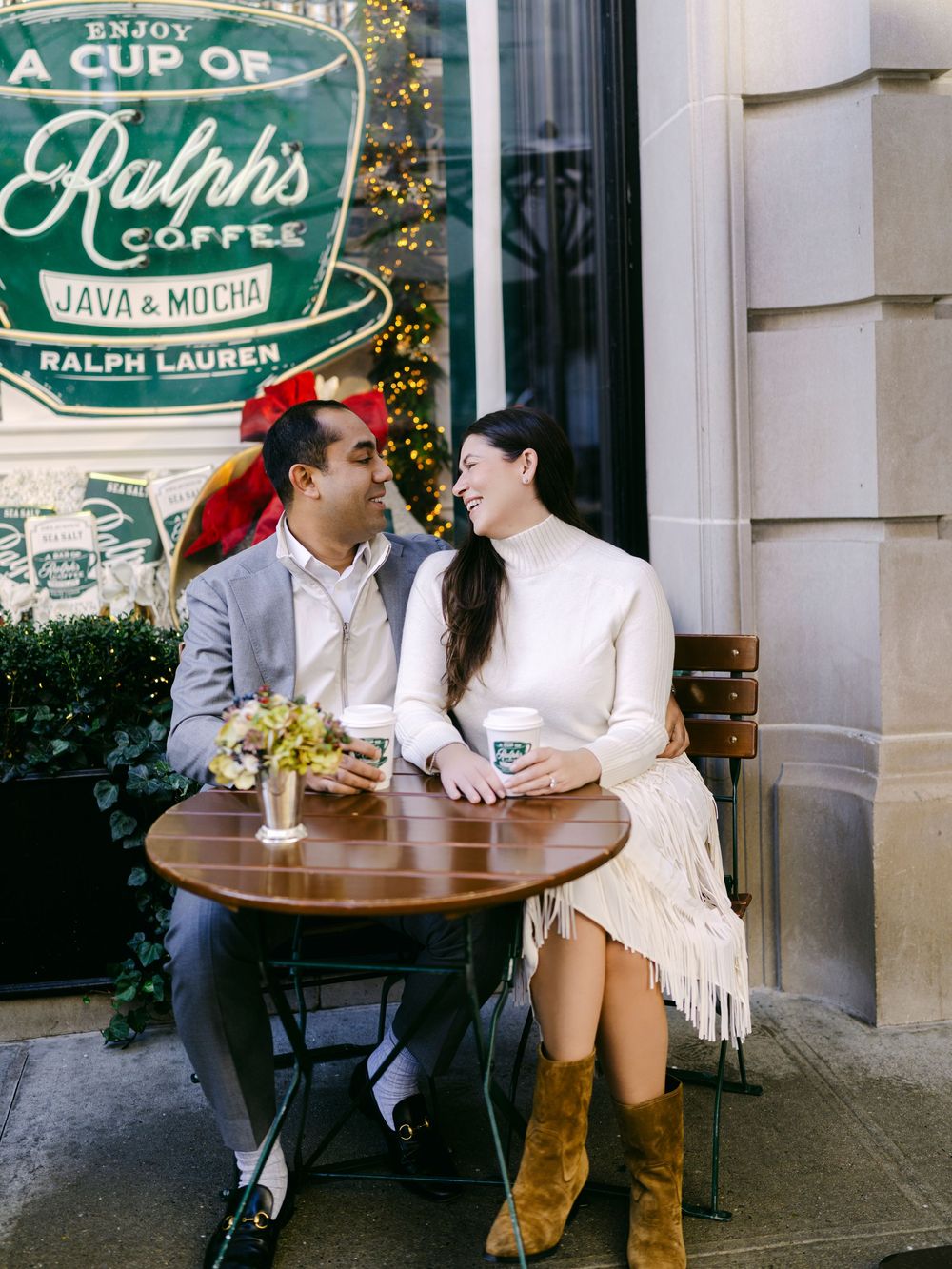 A romantic couple sits at an outdoor cafe table with drinks and flowers, enjoying a warm day together.