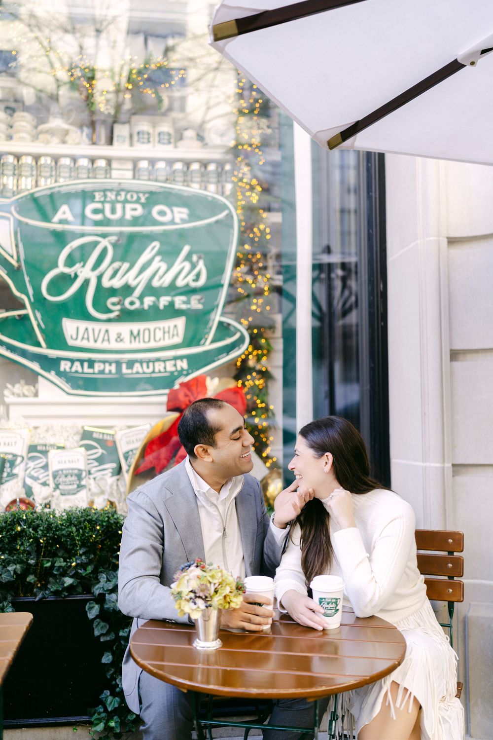 Couple sharing coffee and conversation at an outdoor cafe table with green signage and foliage in background.