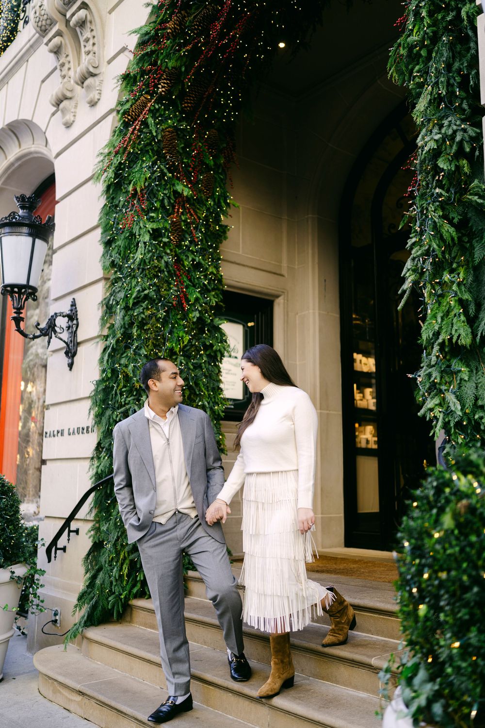 A couple stands on elegant stone steps of a historic building entrance adorned with greenery and holiday decorations.