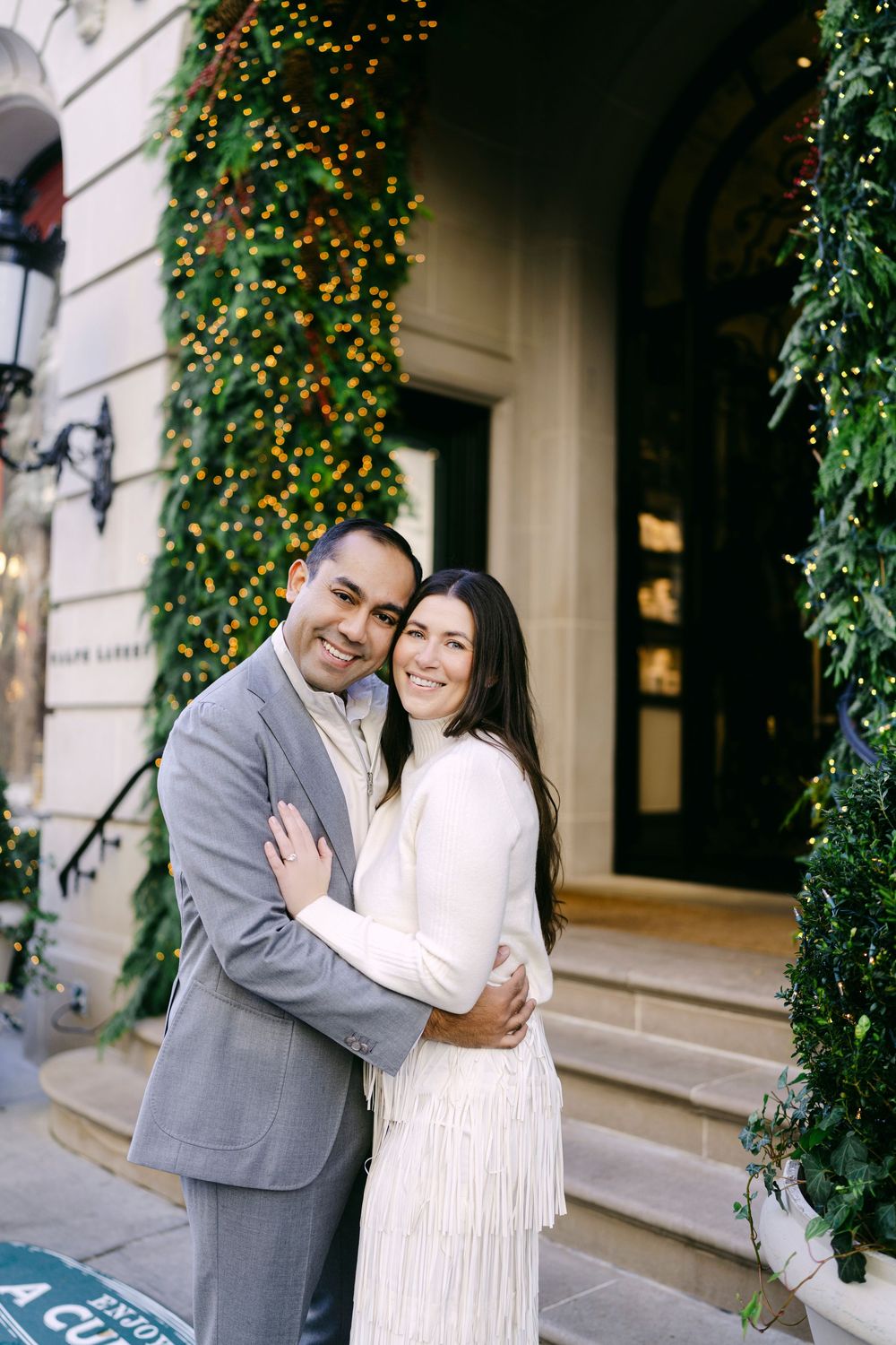 A romantic couple embraces and smiles in front of an elegant building entrance decorated with twinkling lights.