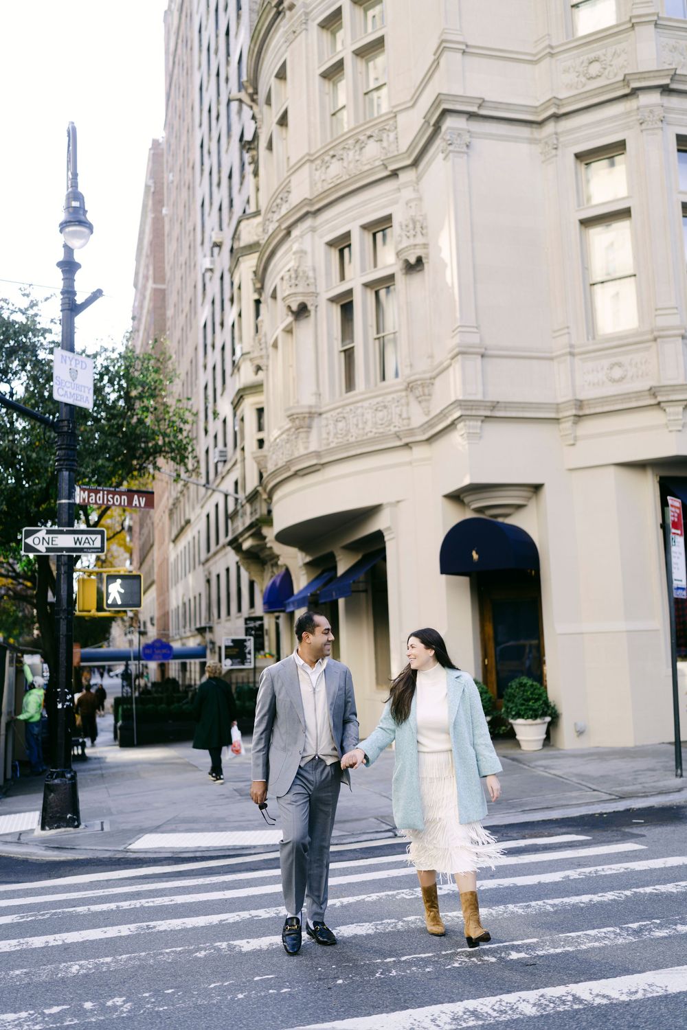 A couple in grey and white outfits walks together on a city street with classic architecture in the background.