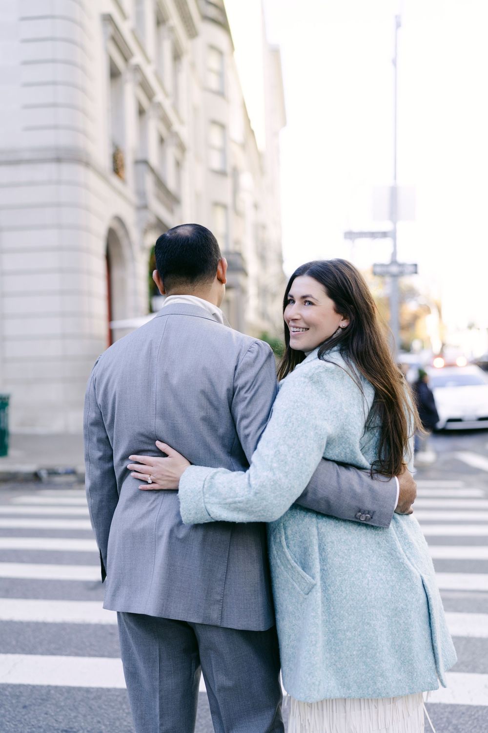 A couple in winter attire embraces while walking along an urban street with classic architecture.