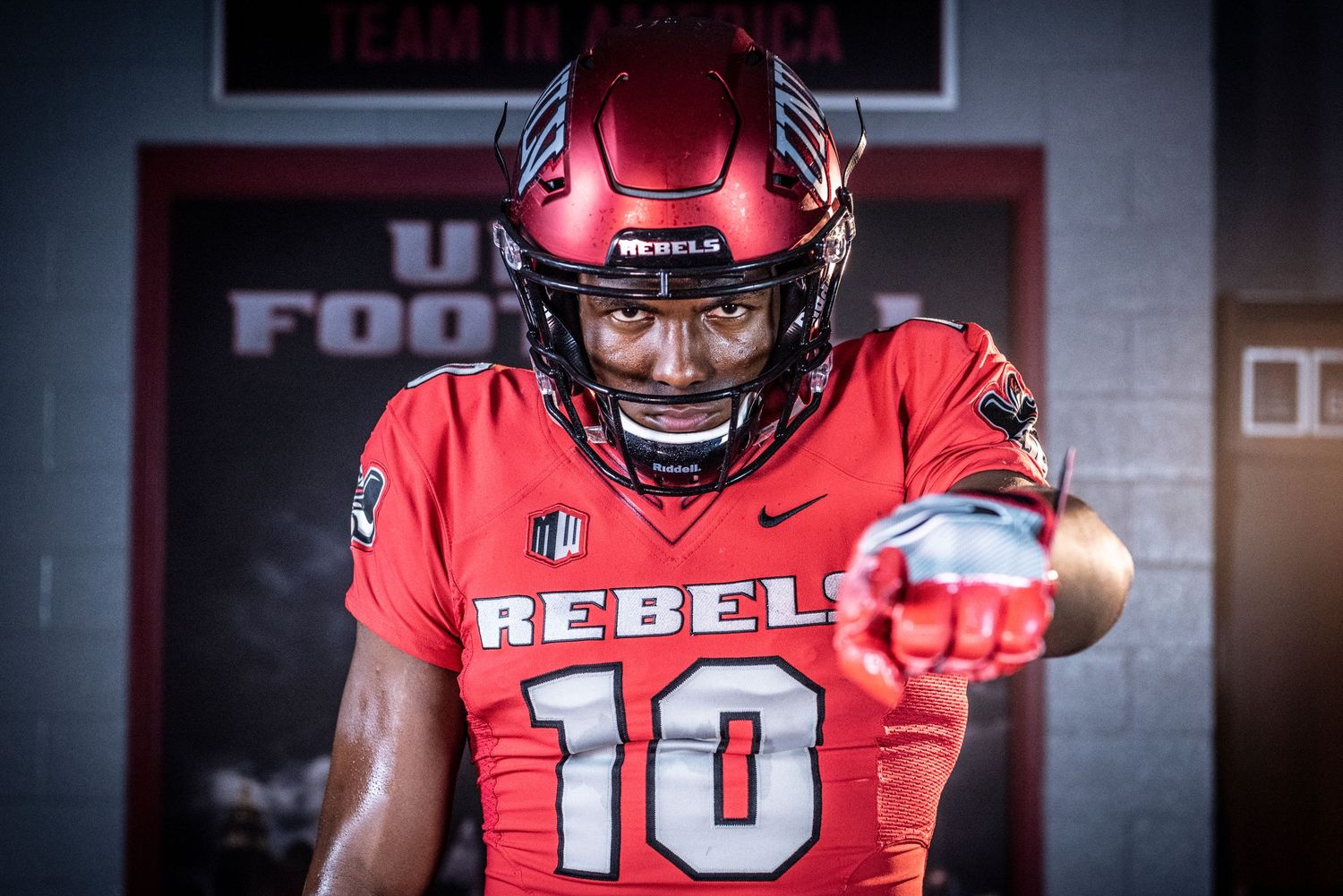 Football player in red UNLV Rebels jersey and helmet poses dramatically in dark locker room.