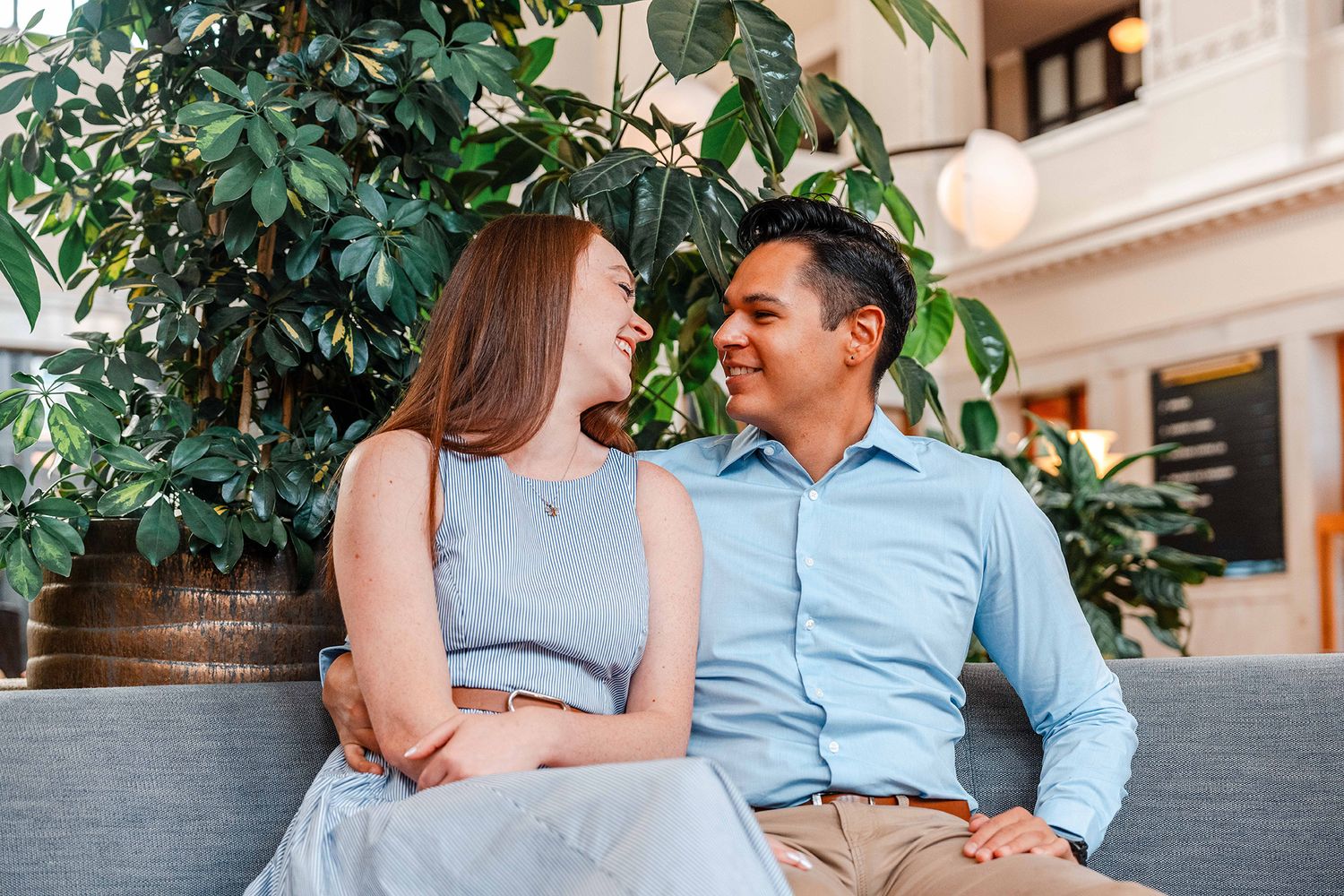 A happy couple laughs and smiles at each other while sitting together on a couch near lush green plants.
