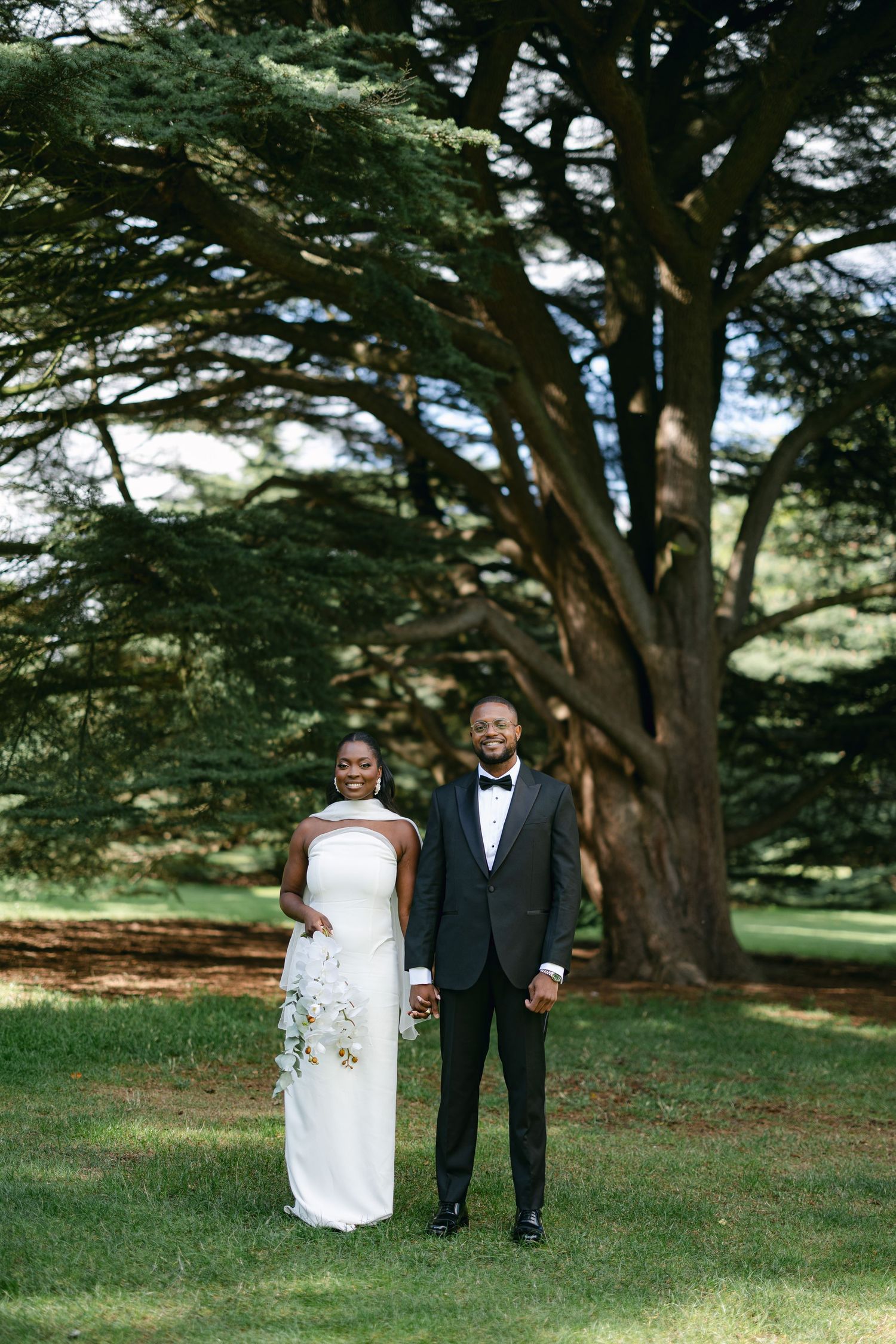 A couple in formal wedding attire stands together under majestic cypress trees in a lush green garden setting.