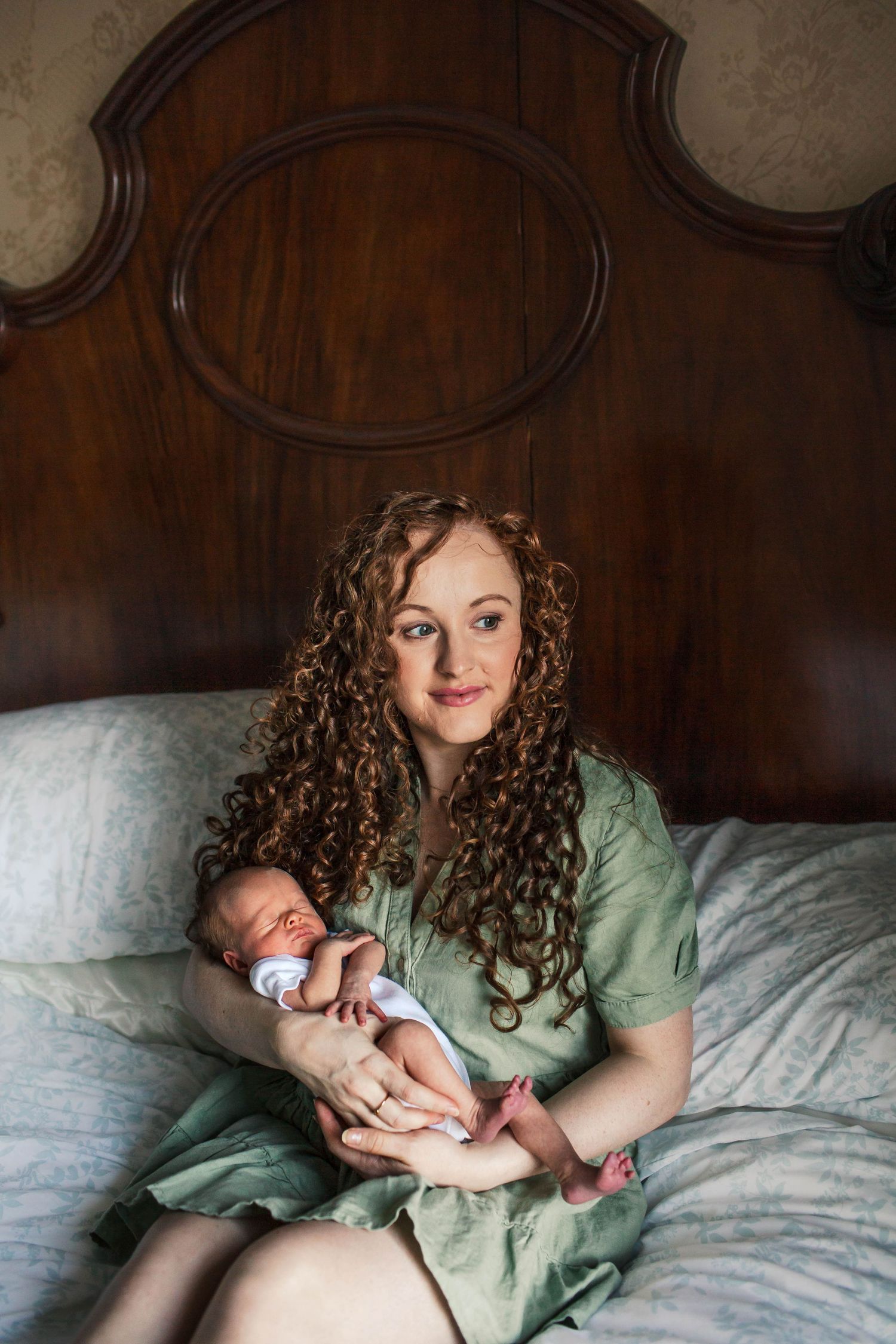 A woman in a mint green dress tenderly holds and kisses a newborn baby wrapped in a white blanket.
