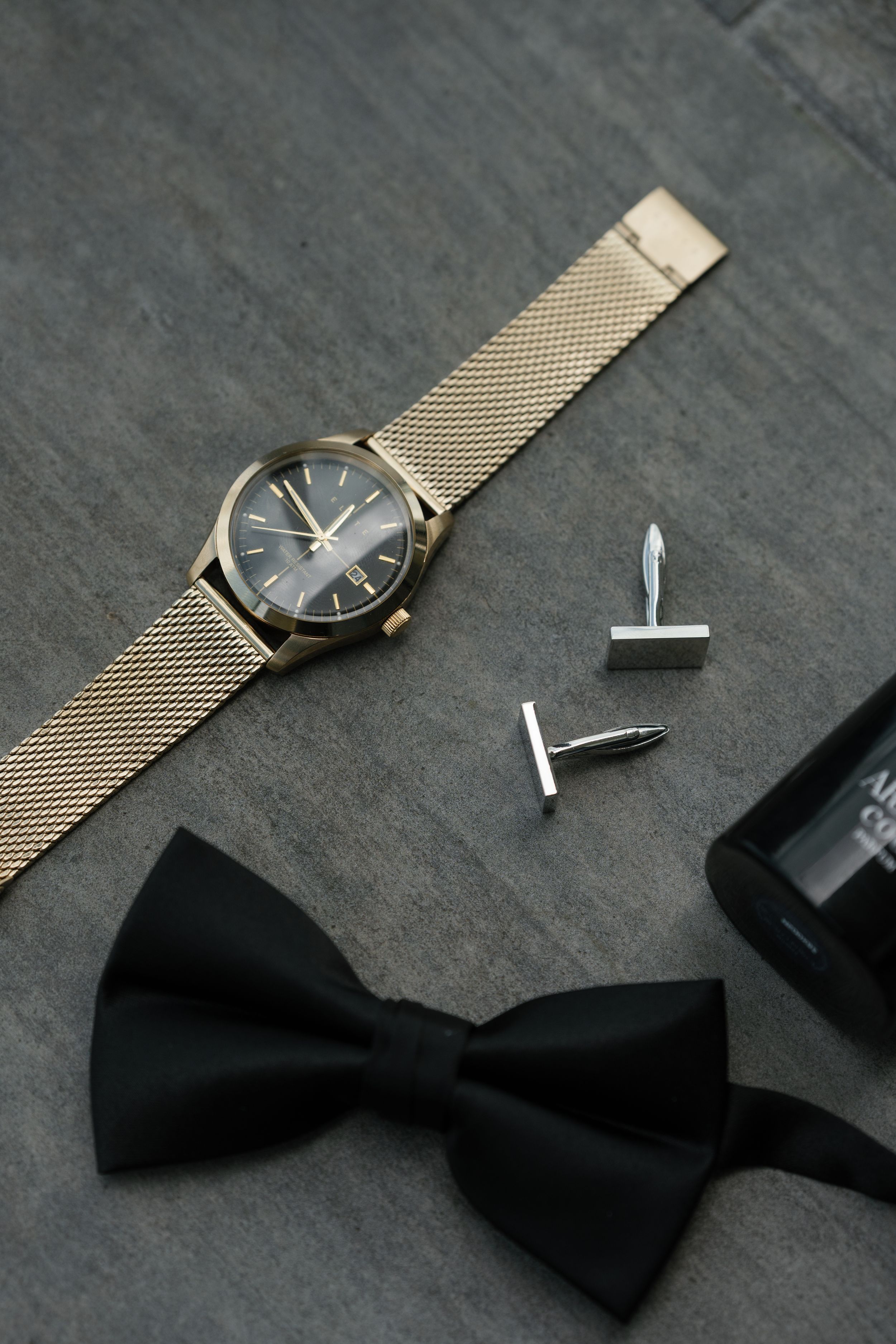 Elegant gold mesh watch with black dial displayed next to a black bow tie and silver cufflinks on dark surface.