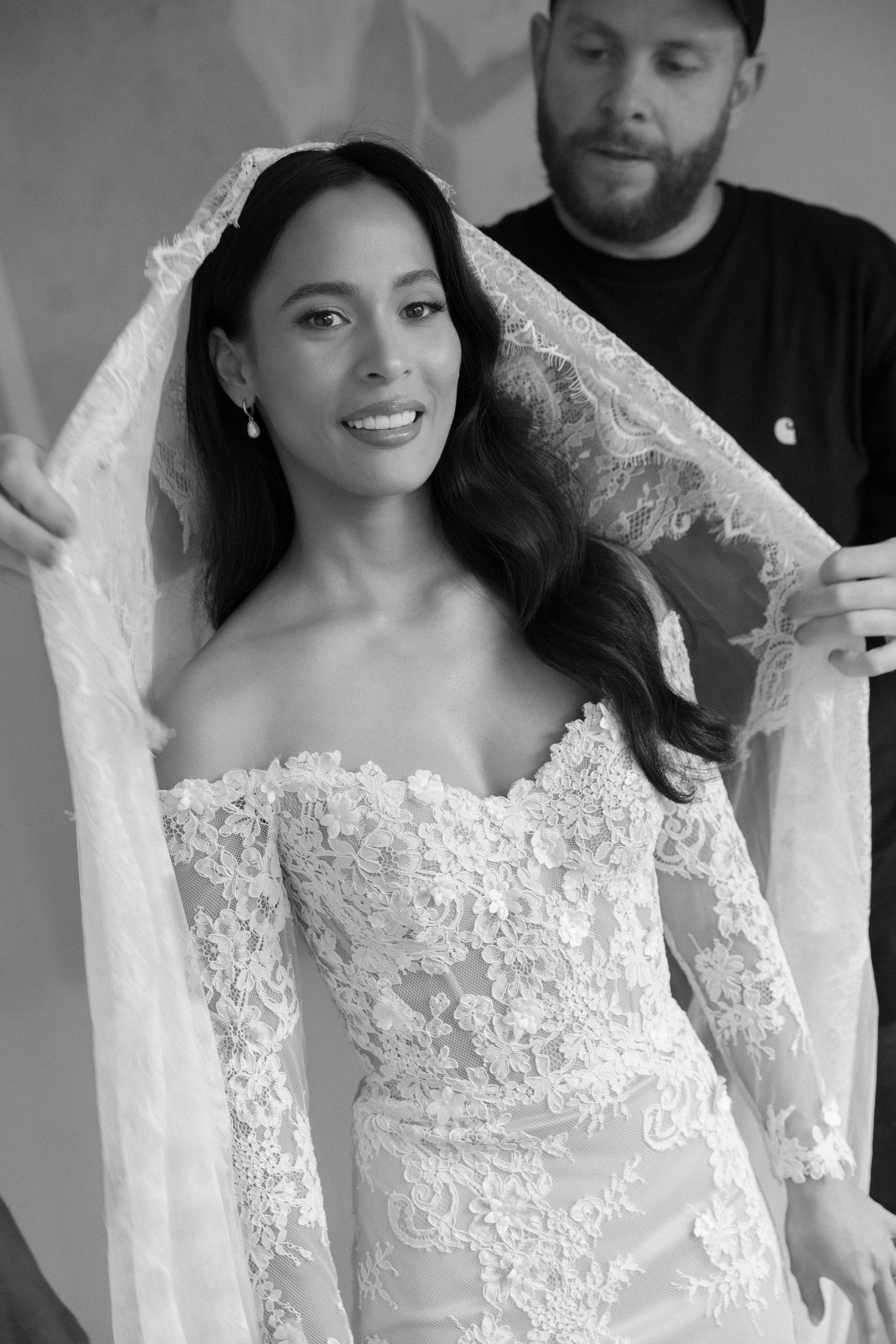 Black and white portrait of a bride wearing an elegant off-shoulder lace wedding gown with long sleeves and veil.