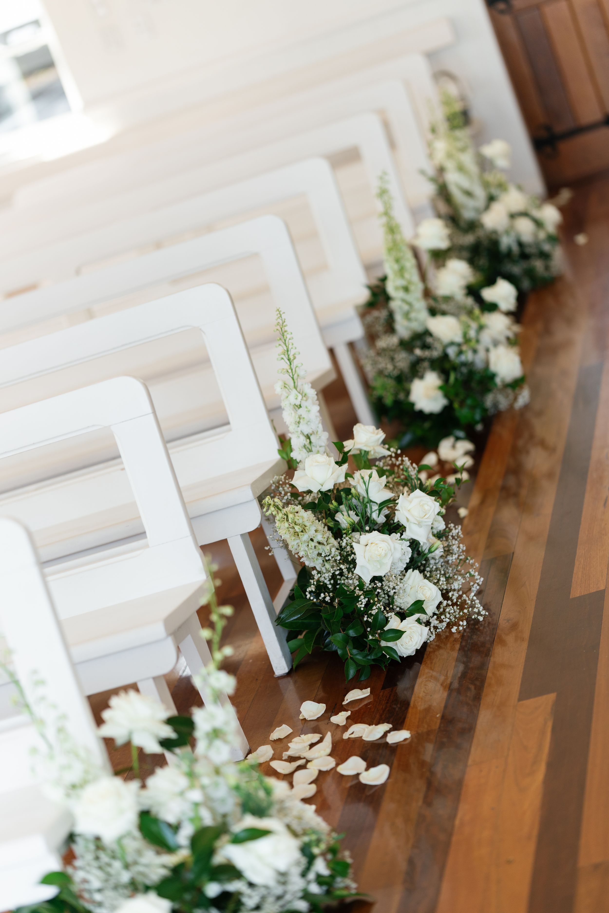 White floral arrangements line a wooden church aisle with elegant pew decorations.