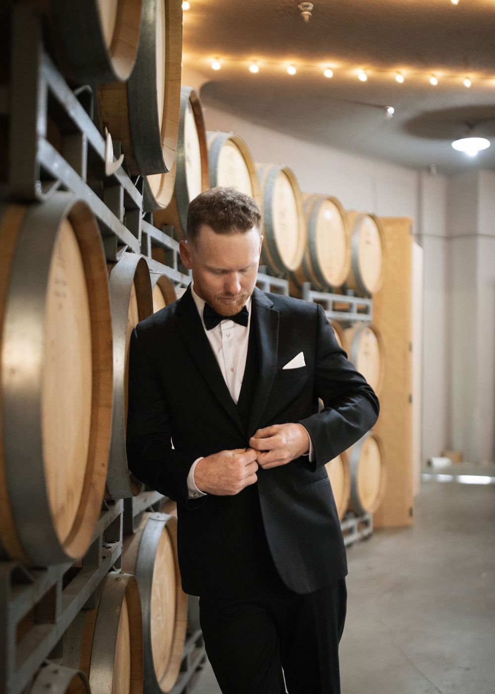 Elegant wine cellar with wooden barrels lining the walls and a person in formal attire adjusting their outfit.