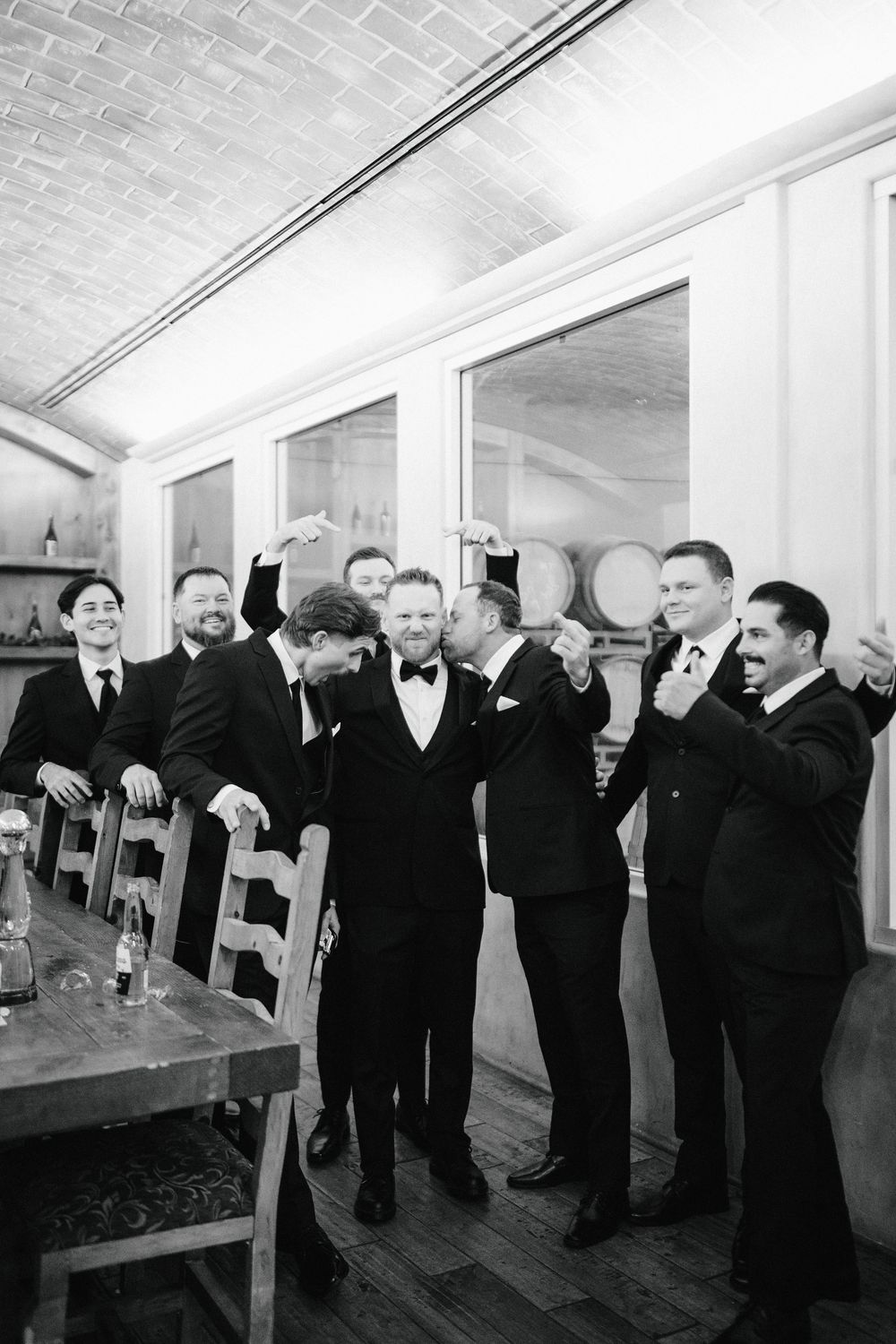 A group of formally dressed men in black suits and tuxedos stand together in an elegant indoor setting.