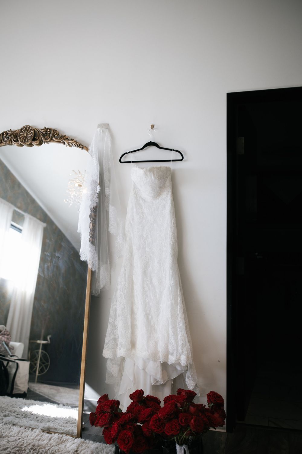 A delicate white lace wedding dress hangs against a light wall with red roses placed below.