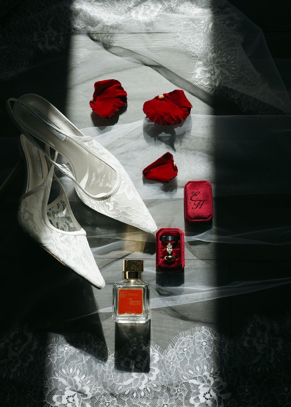 Wedding accessories including white shoes, red rose petals, and perfume bottles arranged on a reflective surface.