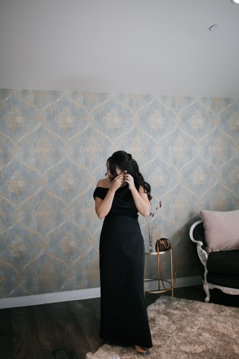 Person in long black dress standing against patterned wallpaper in elegant room setting.