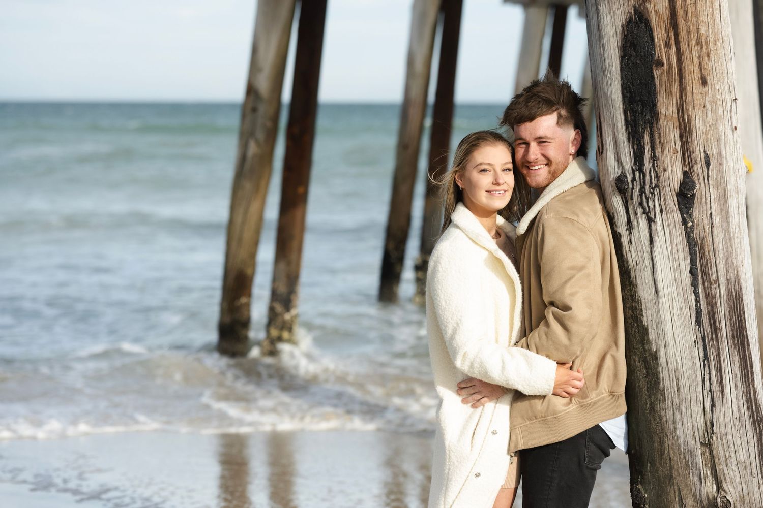 Couple embracing near a beach pier, smiling warmly in casual attire with ocean waves in the background.