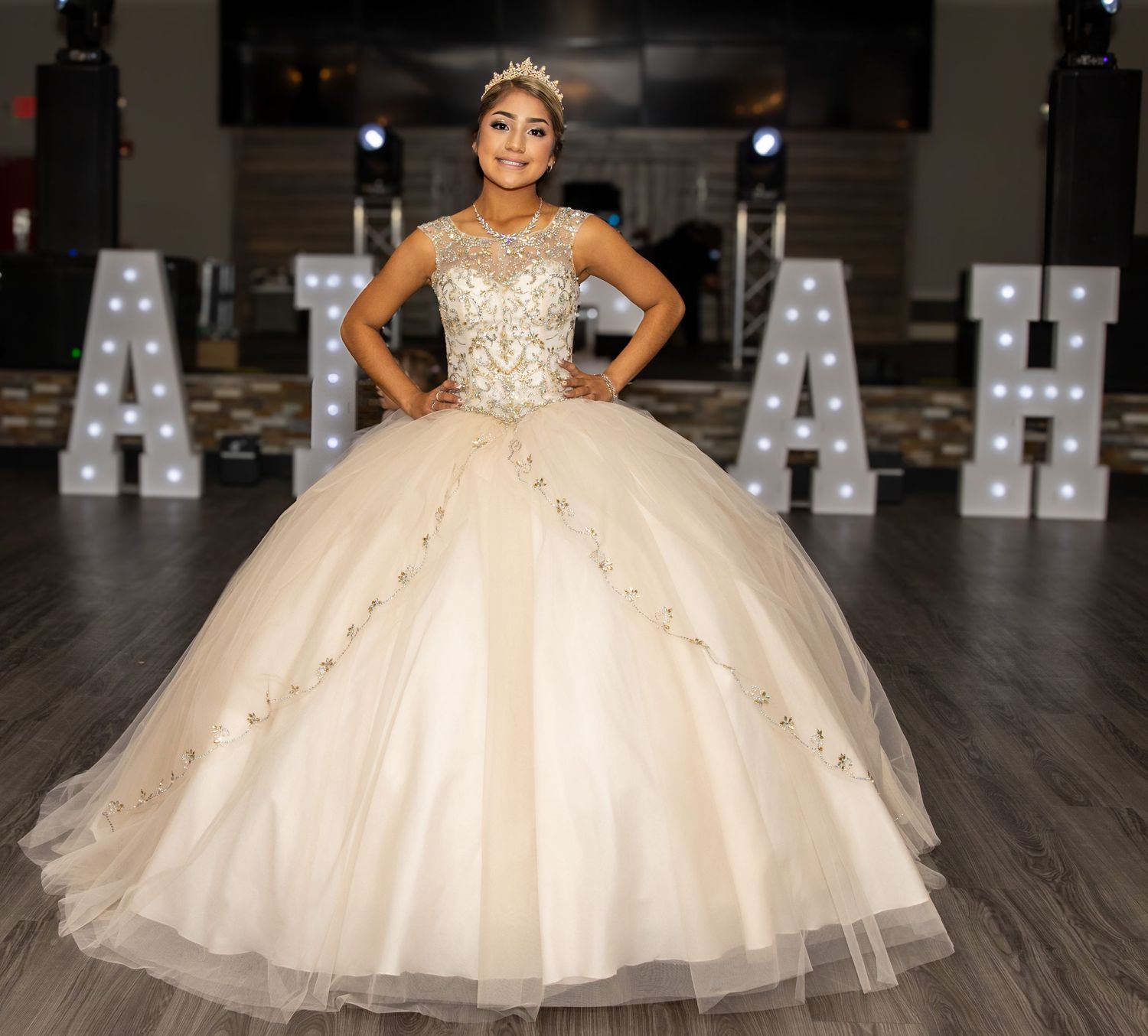 A quinceañera dress in champagne color with crystal embellishments and a full tulle ballgown skirt sparkles in dim lighting.