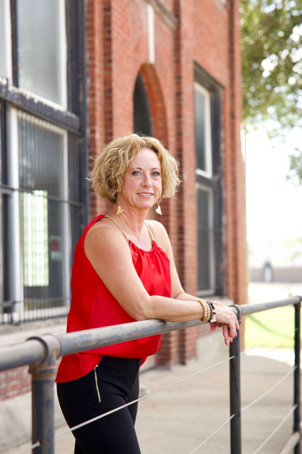 A person in a red sleeveless top and dark pants leans against a railing outside a brick building.