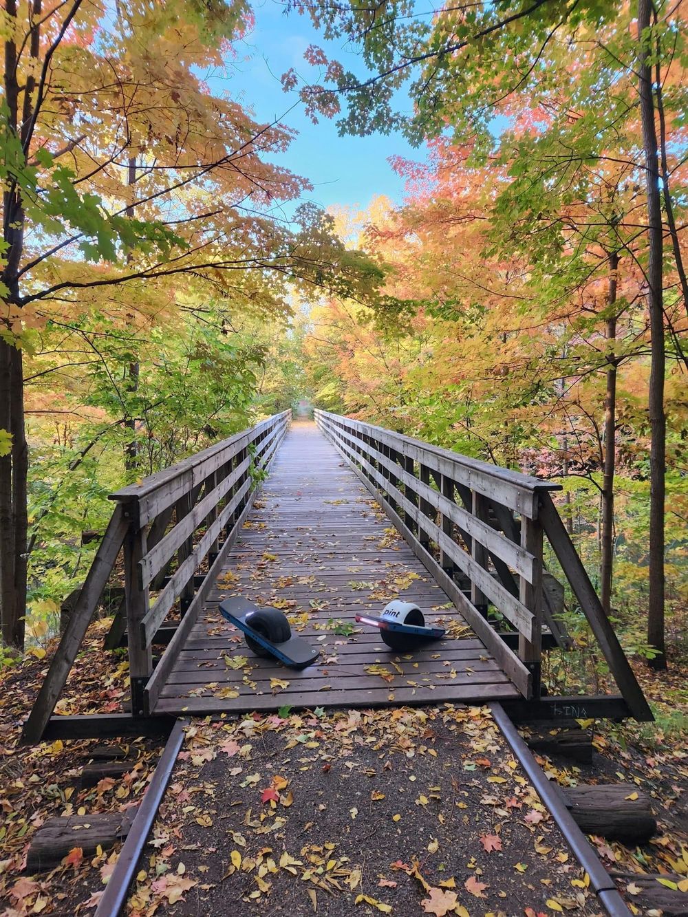 A wooden footbridge surrounded by colorful autumn foliage with fallen leaves scattered across its planks.
