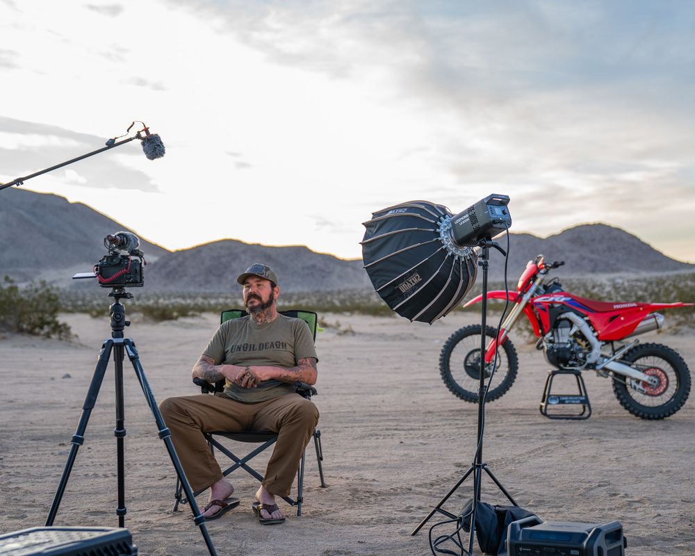 Photography equipment and motorcycle set up in a desert landscape during sunset with lighting gear and camera rigs.