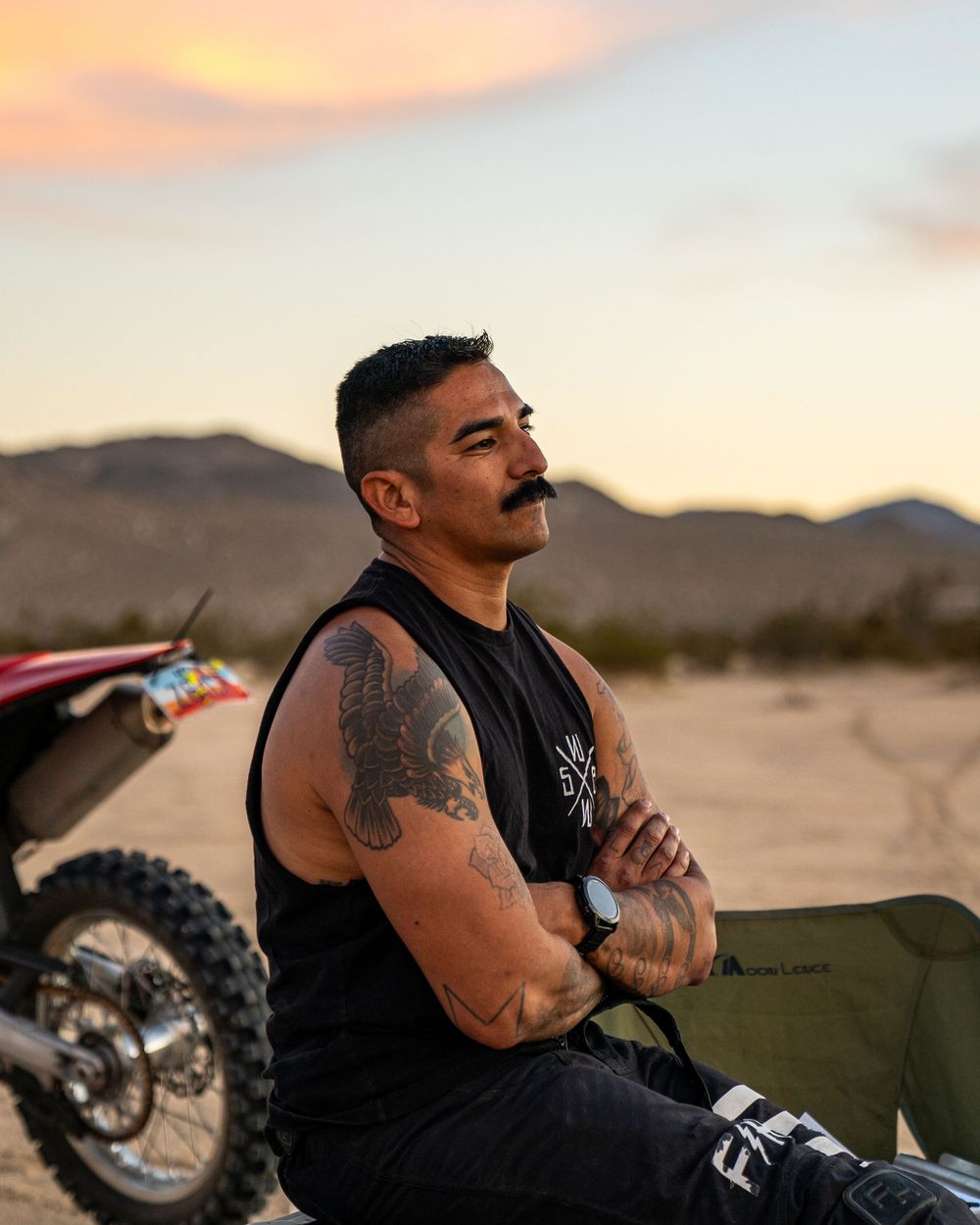 Motorcyclist in black tank top sits on bike against desert landscape at sunset with mountains in background.