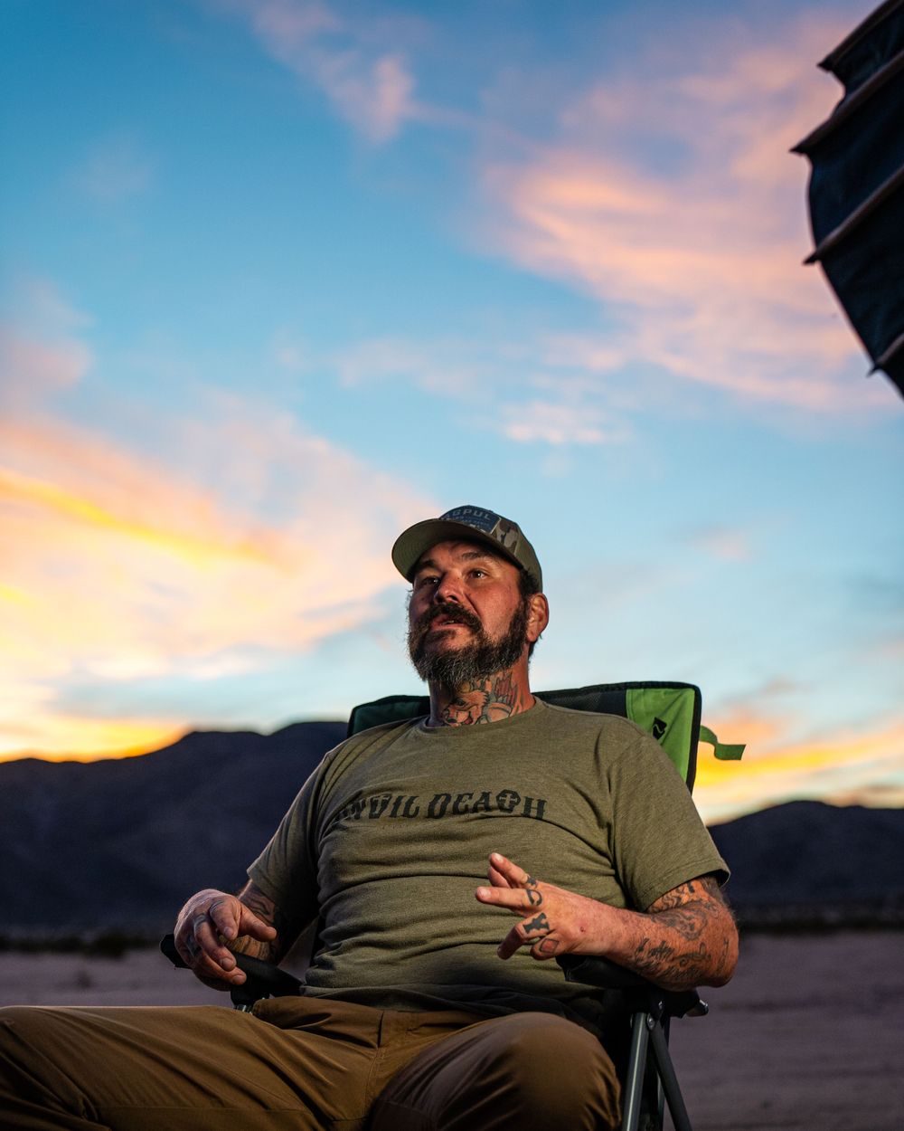 Person in olive green t-shirt sits in camping chair against vibrant sunset sky and mountain backdrop.
