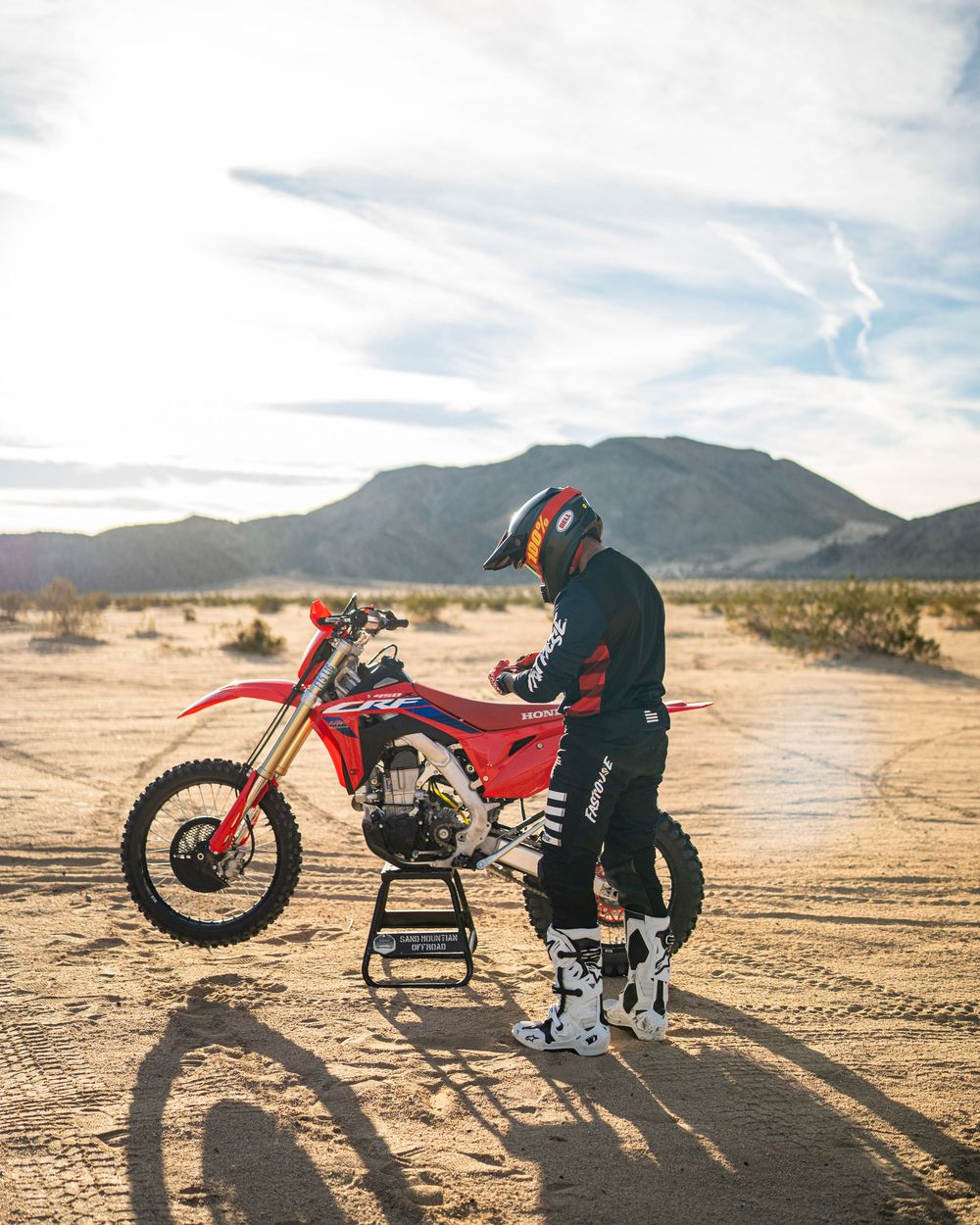 A red dirt bike and rider cast long shadows on desert terrain with mountains in the background at sunset.