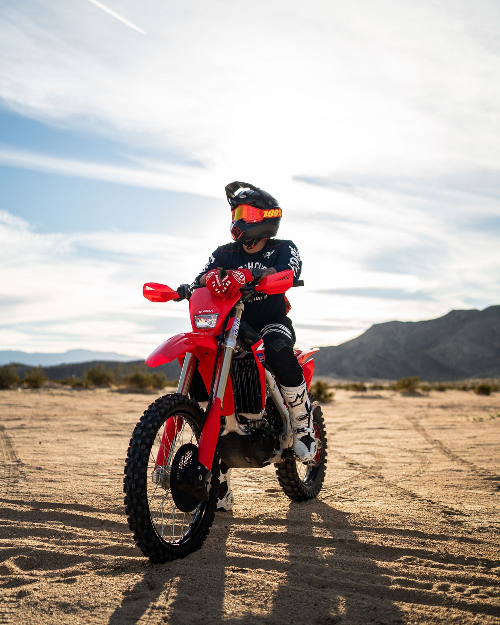 Red dirt bike parked in desert terrain with mountains in background during sunset casting long shadows.