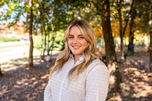 Someone in a cozy white sweater smiles warmly while standing in a sunlit autumn forest setting.