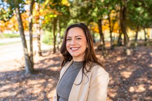 Person wearing a gray sweater smiles while sitting outdoors among autumn trees with golden foliage.