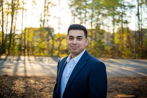 Professional portrait of someone in a navy suit and light blue shirt standing outdoors with sunlit autumn trees in background.