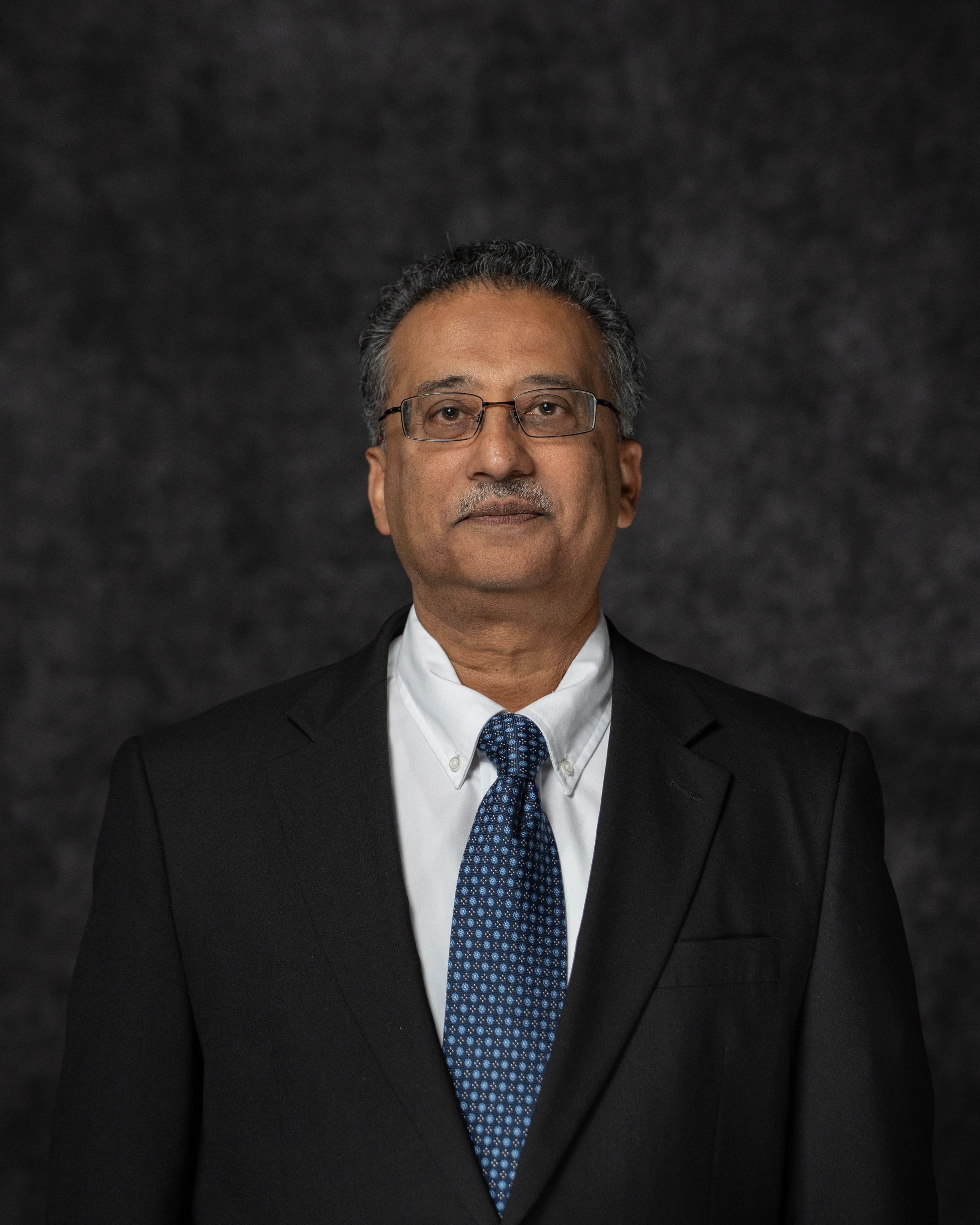 Professional headshot of a middle-aged man wearing glasses, dark suit, white shirt, and blue tie against gray backdrop.