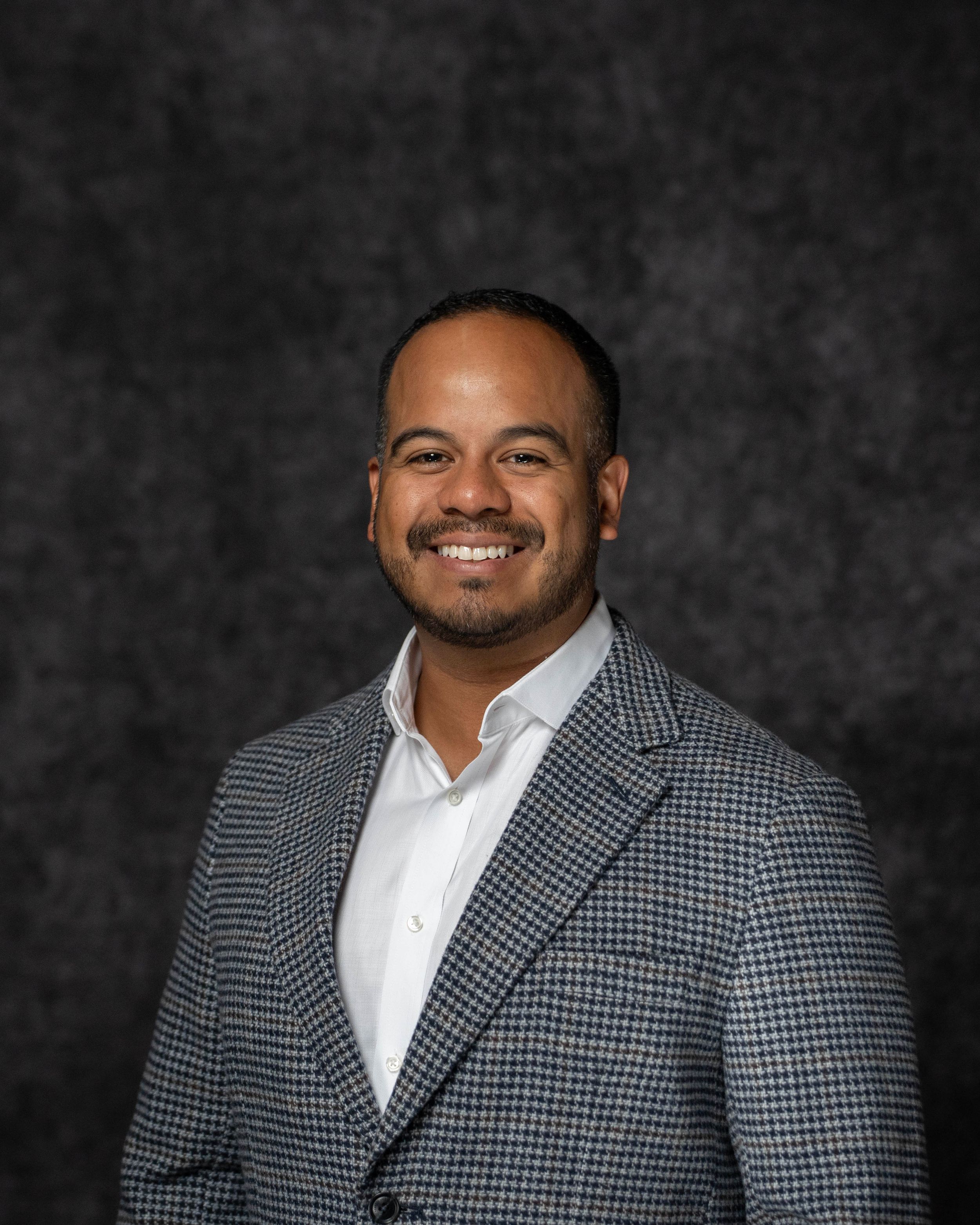 Professional headshot of a smiling man in a gray plaid blazer and white shirt against a dark background.