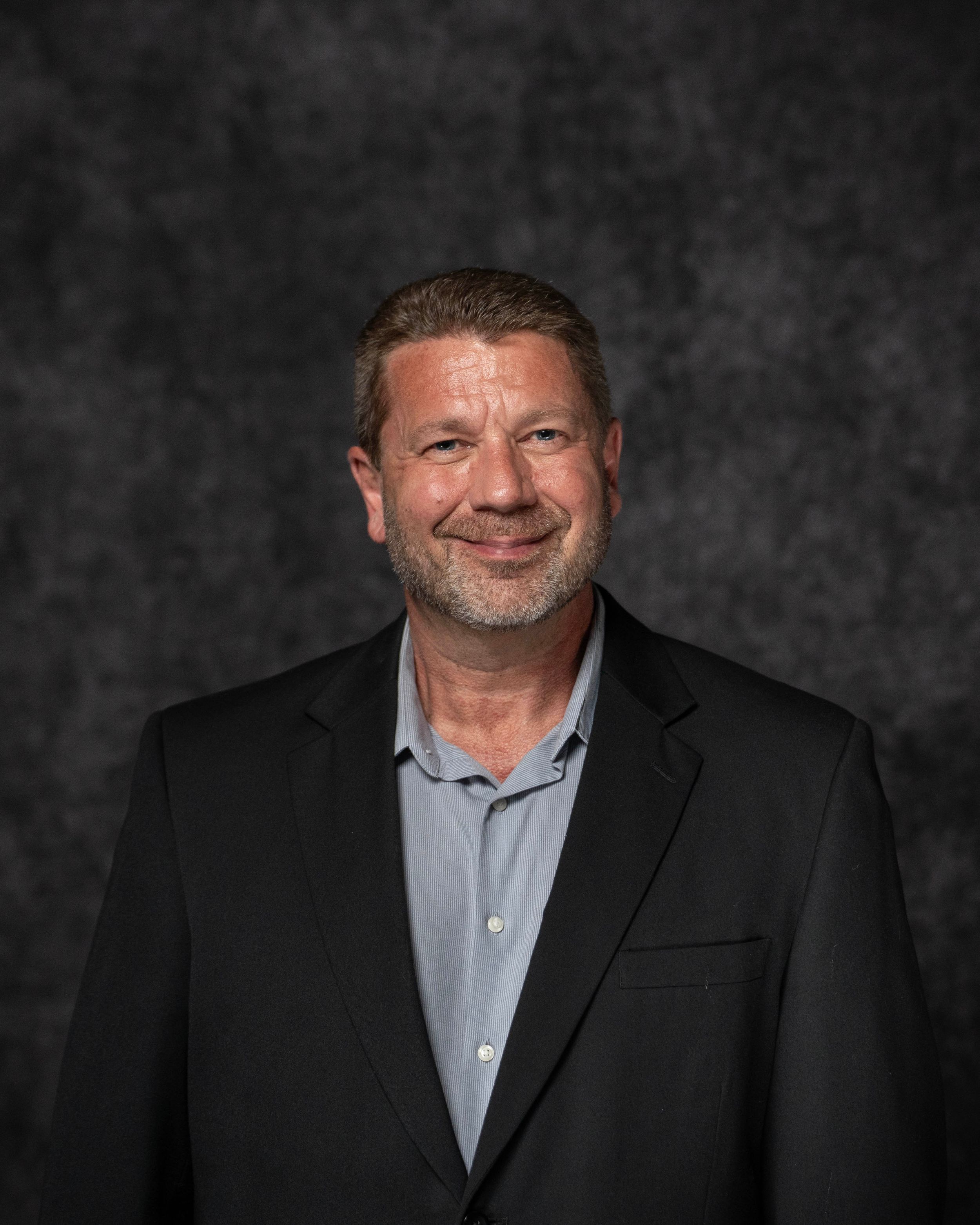Professional headshot of a middle-aged man with short hair and beard wearing a black blazer and striped shirt.