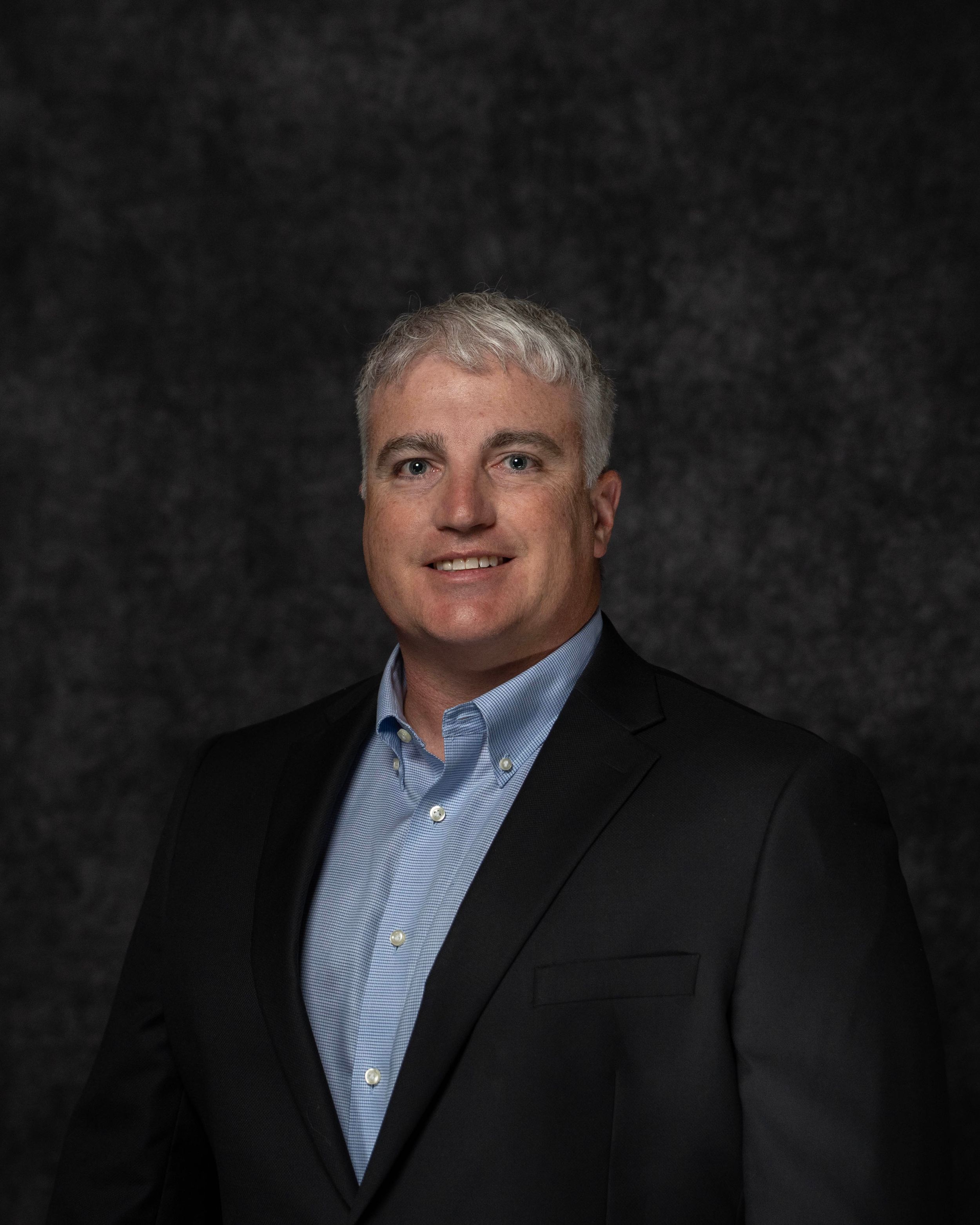 Professional headshot of a middle-aged man with gray hair wearing a black blazer and light blue shirt.