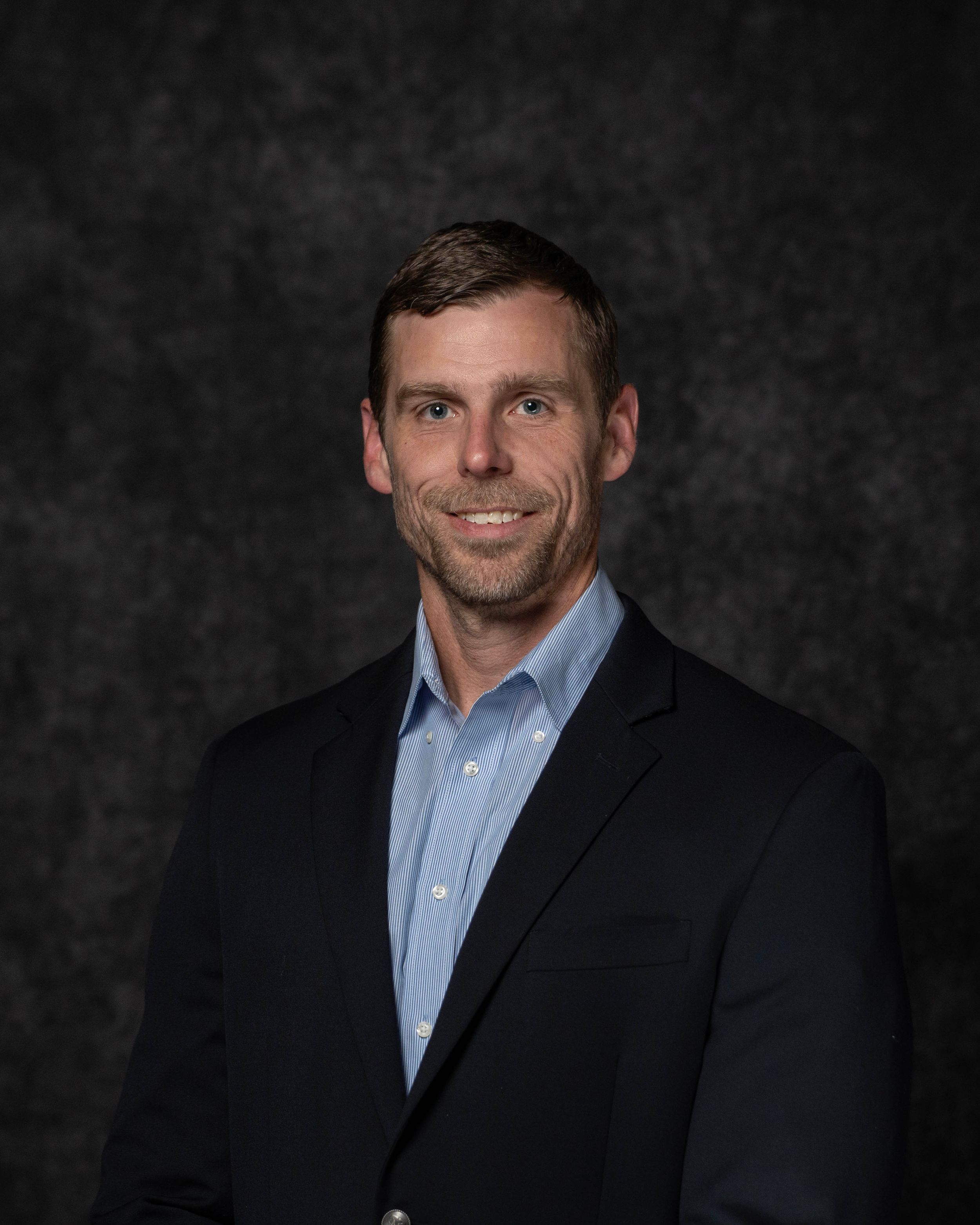 Professional headshot of a smiling man in a dark blazer and light blue shirt against a gray backdrop.