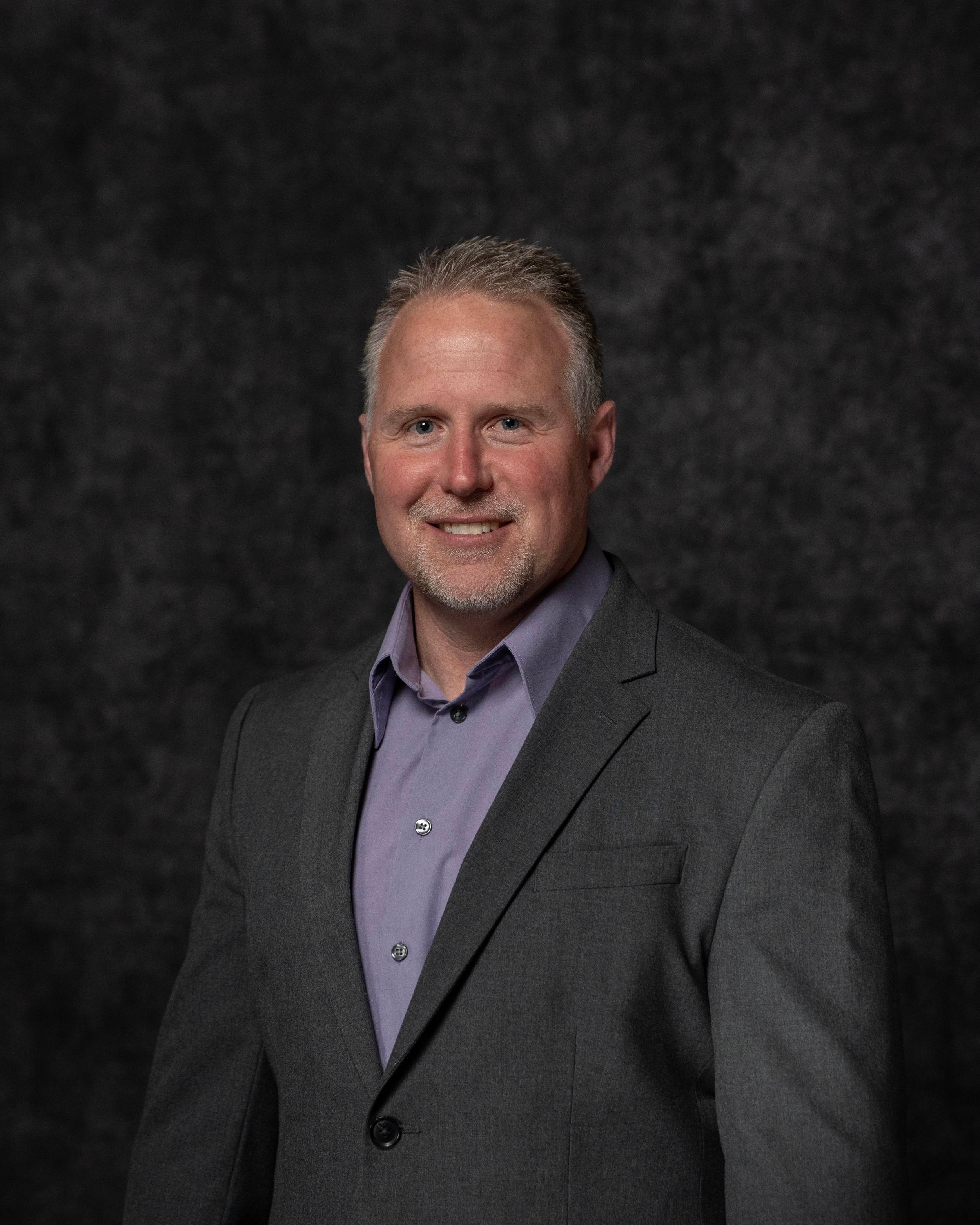 Professional headshot of a smiling middle-aged man with gray hair wearing a gray blazer and purple dress shirt.
