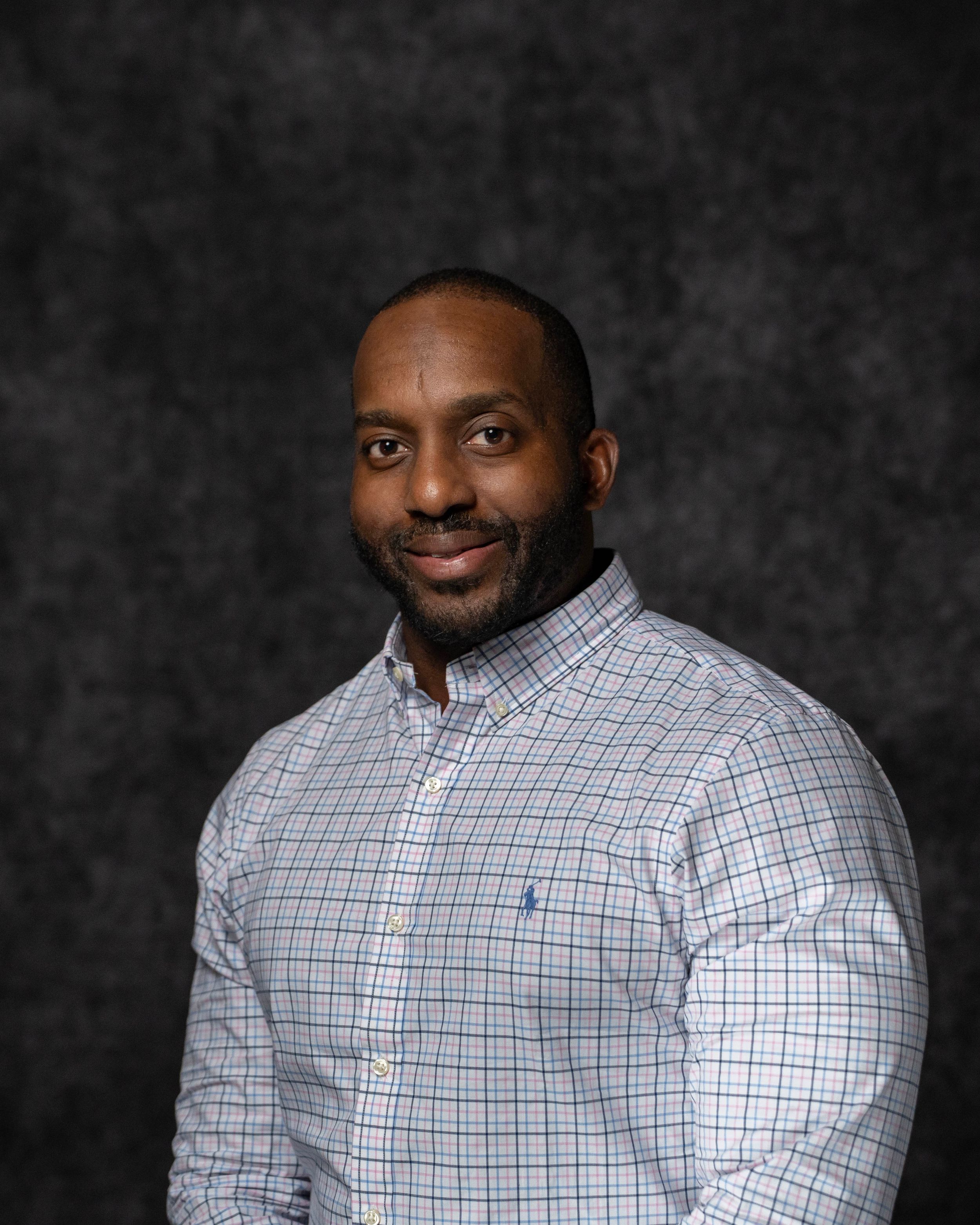 Professional headshot of a smiling man in a plaid button-up shirt against a dark studio background.