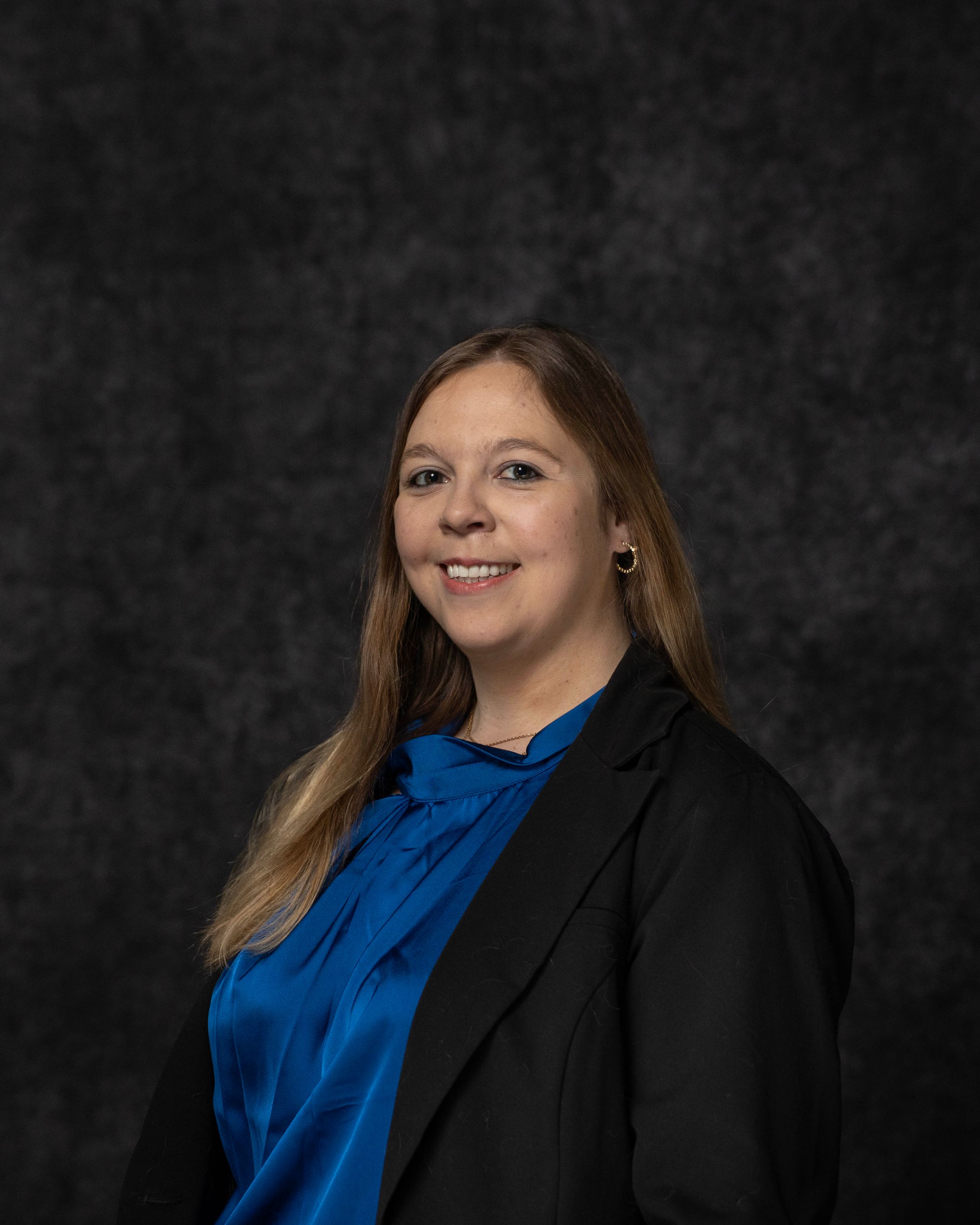 Professional woman in blue blouse and black blazer smiling against dark studio background.