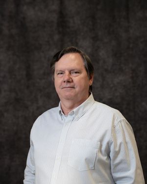 Middle-aged man with dark hair wearing a light blue striped button-up shirt, posing for a professional headshot.