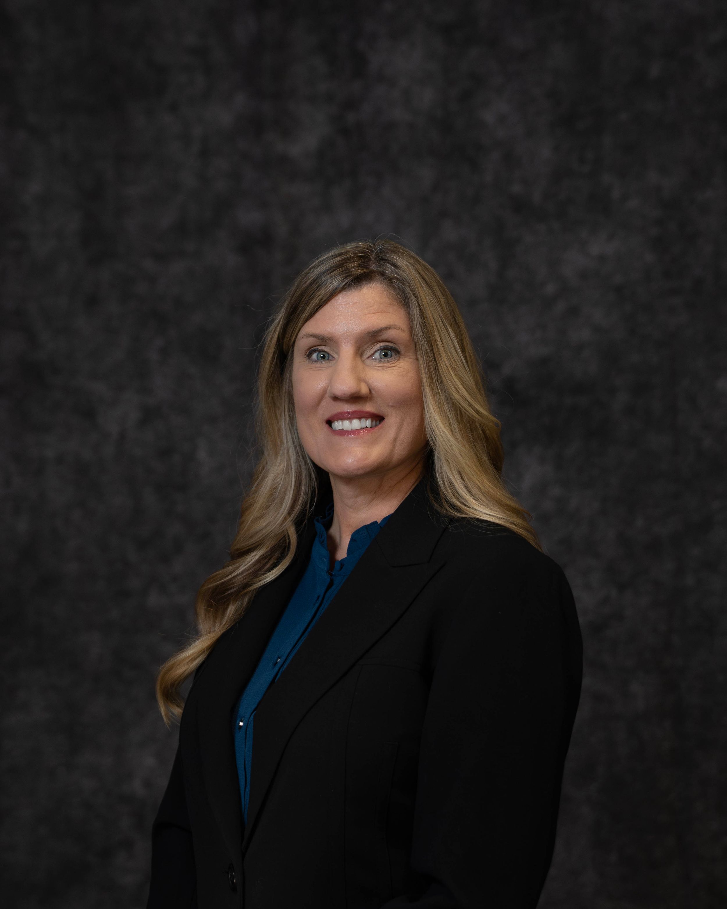 Professional woman in black blazer and teal blouse smiling against dark gray studio background.