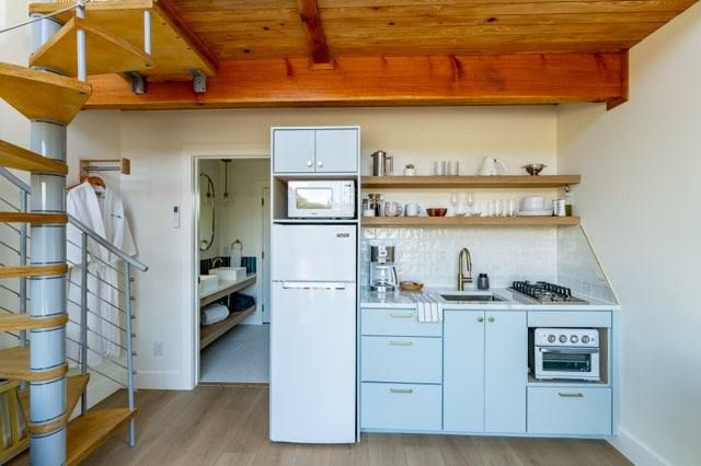 Modern compact kitchen with light blue cabinets, white refrigerator, and floating wooden shelves in a studio apartment.