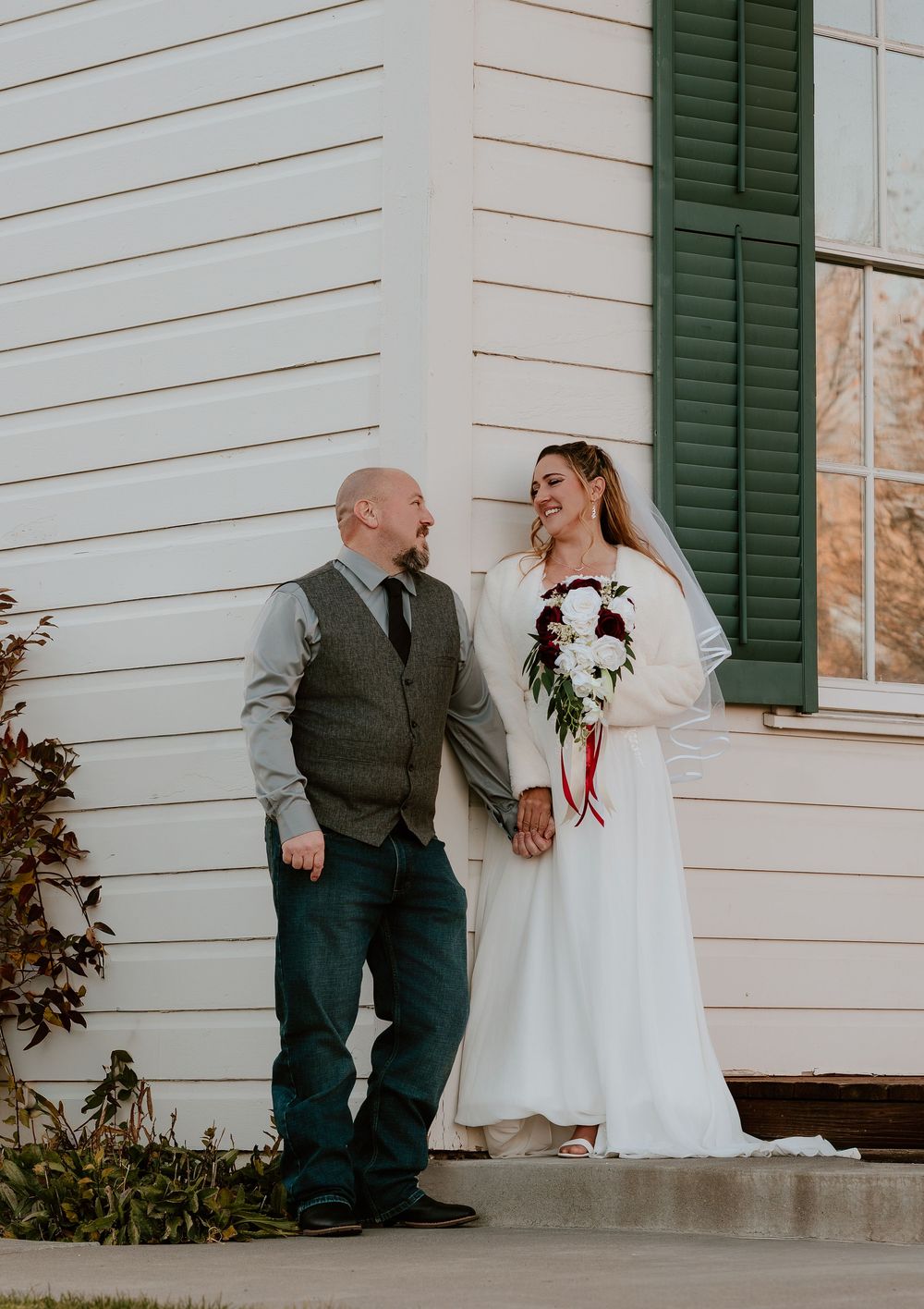 A wedding couple sits together on white porch steps with green shutters in the background.