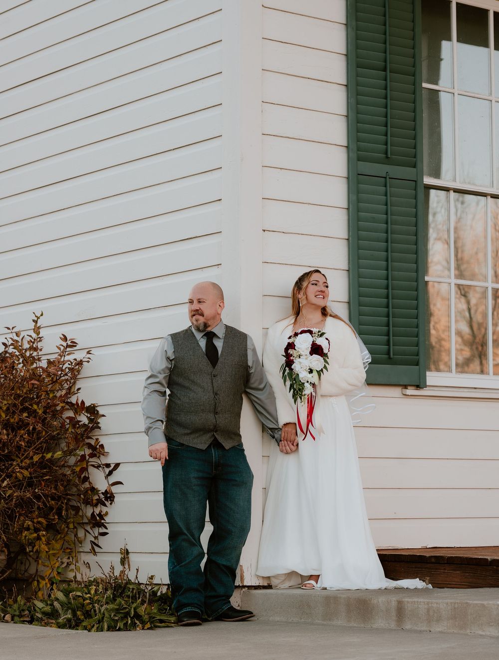 A bride and groom share a candid moment on a white porch with green shutters during their wedding day.