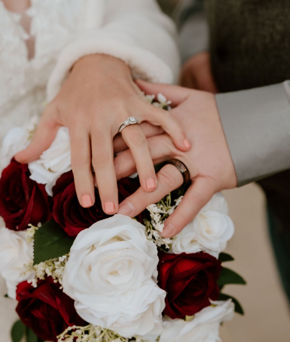 Close-up view of hands with wedding rings holding a bouquet of red and white roses with delicate baby's breath flowers.