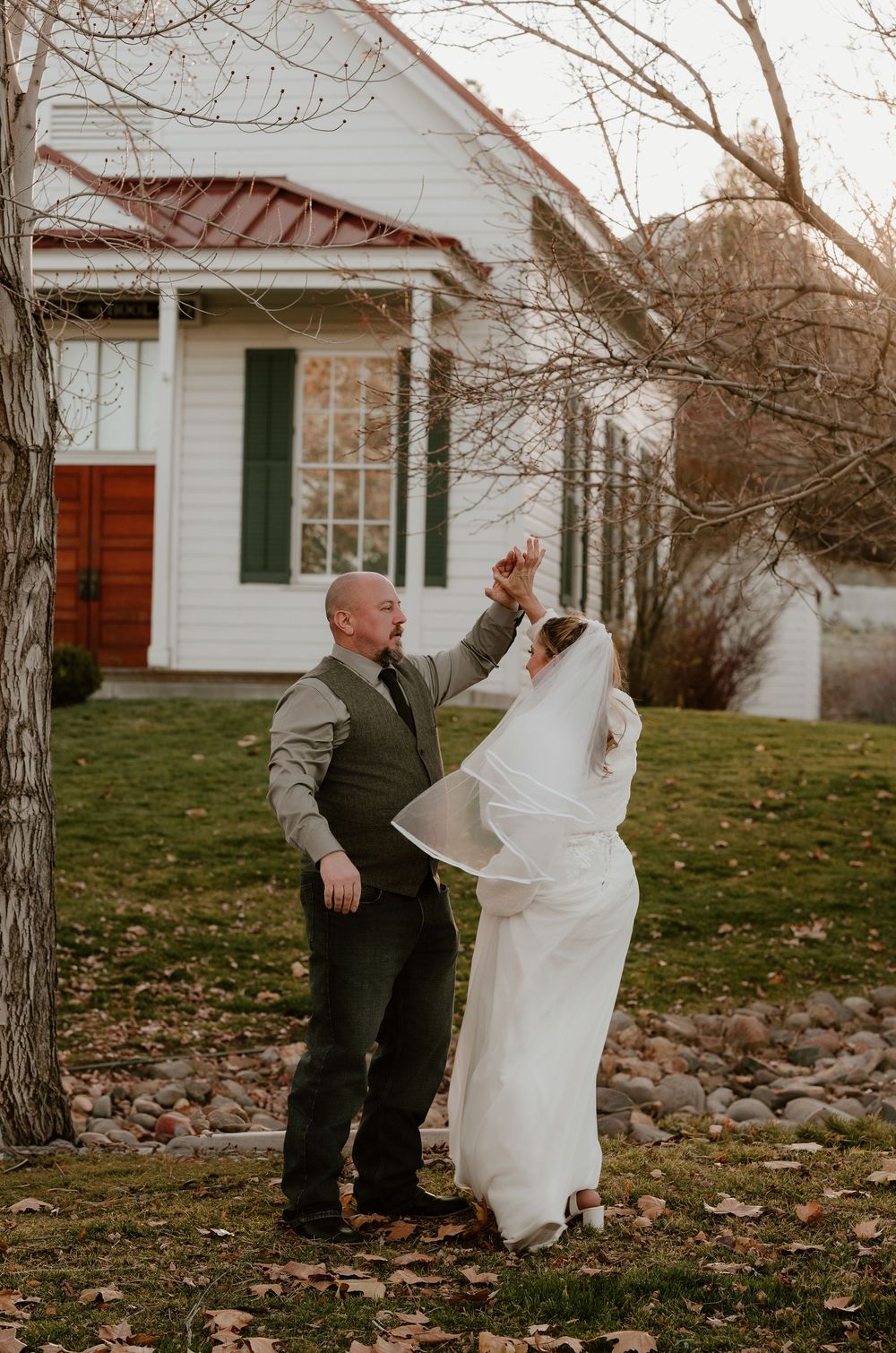 Couple in wedding attire playfully dancing and spinning together in front of a white farmhouse on an autumn day.